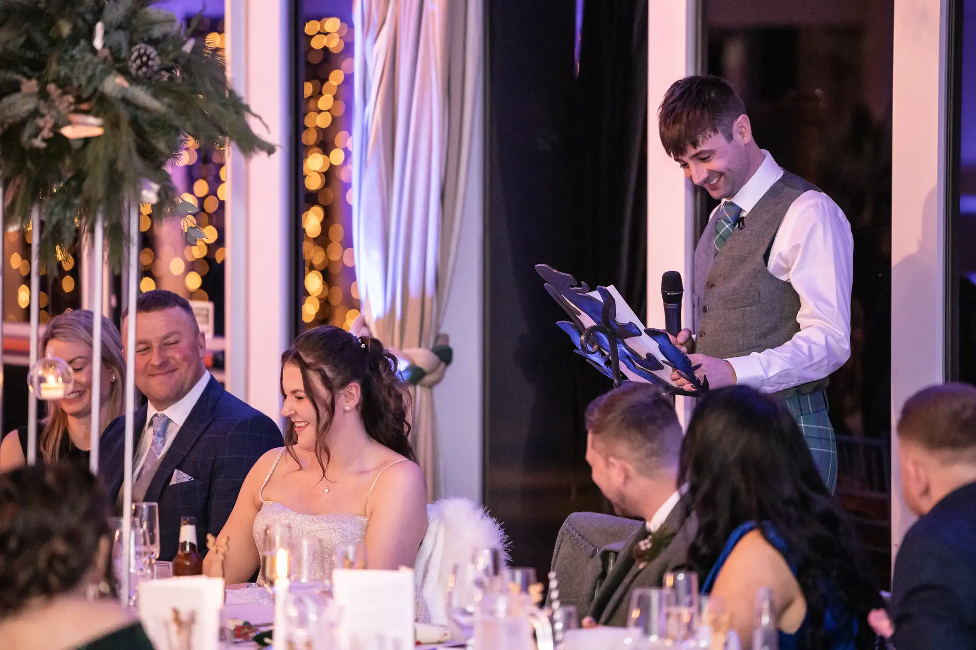 A man in a suit reads from a clipboard while holding a microphone, addressing seated guests at a formal event. The guests are smiling and laughing. A decorated table and lights are visible.