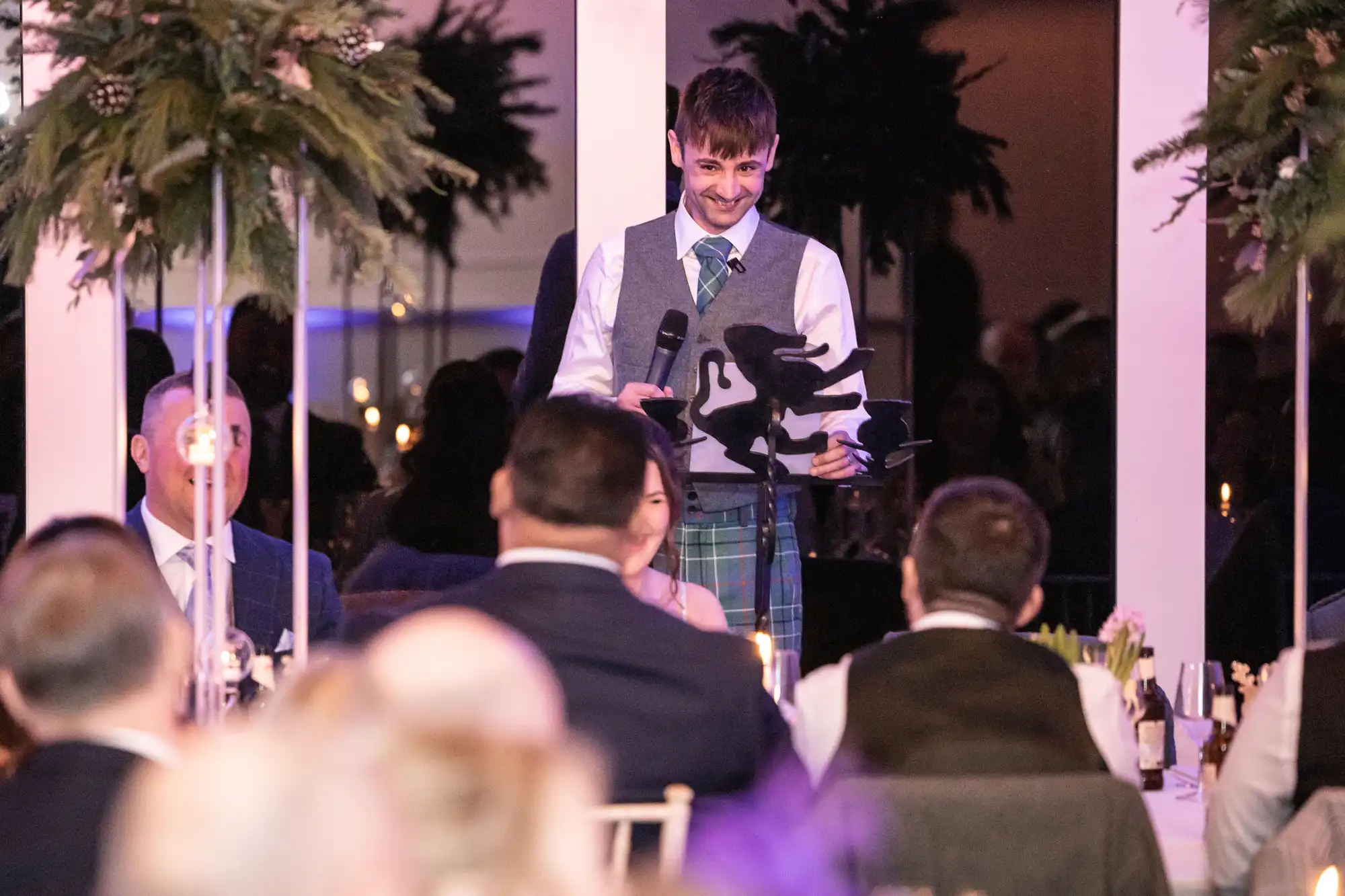 A man in a kilt speaks into a microphone at a formal event, addressing an audience seated at round tables decorated with flowers and greenery.