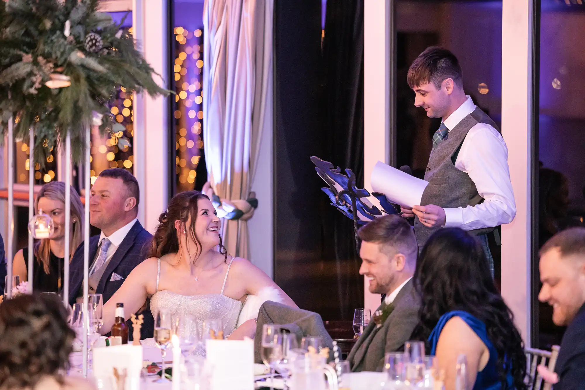 A man in a suit delivers a speech from a podium at a formal event. Guests seated at tables around him listen attentively and smile. The room is decorated with hanging lights and greenery.