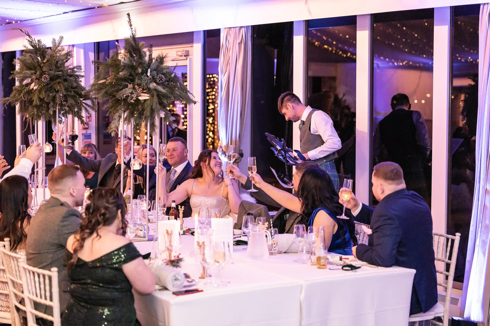 A group of people seated at a decorated table raise their glasses in a toast as a man stands and speaks at a podium in a well-lit venue.