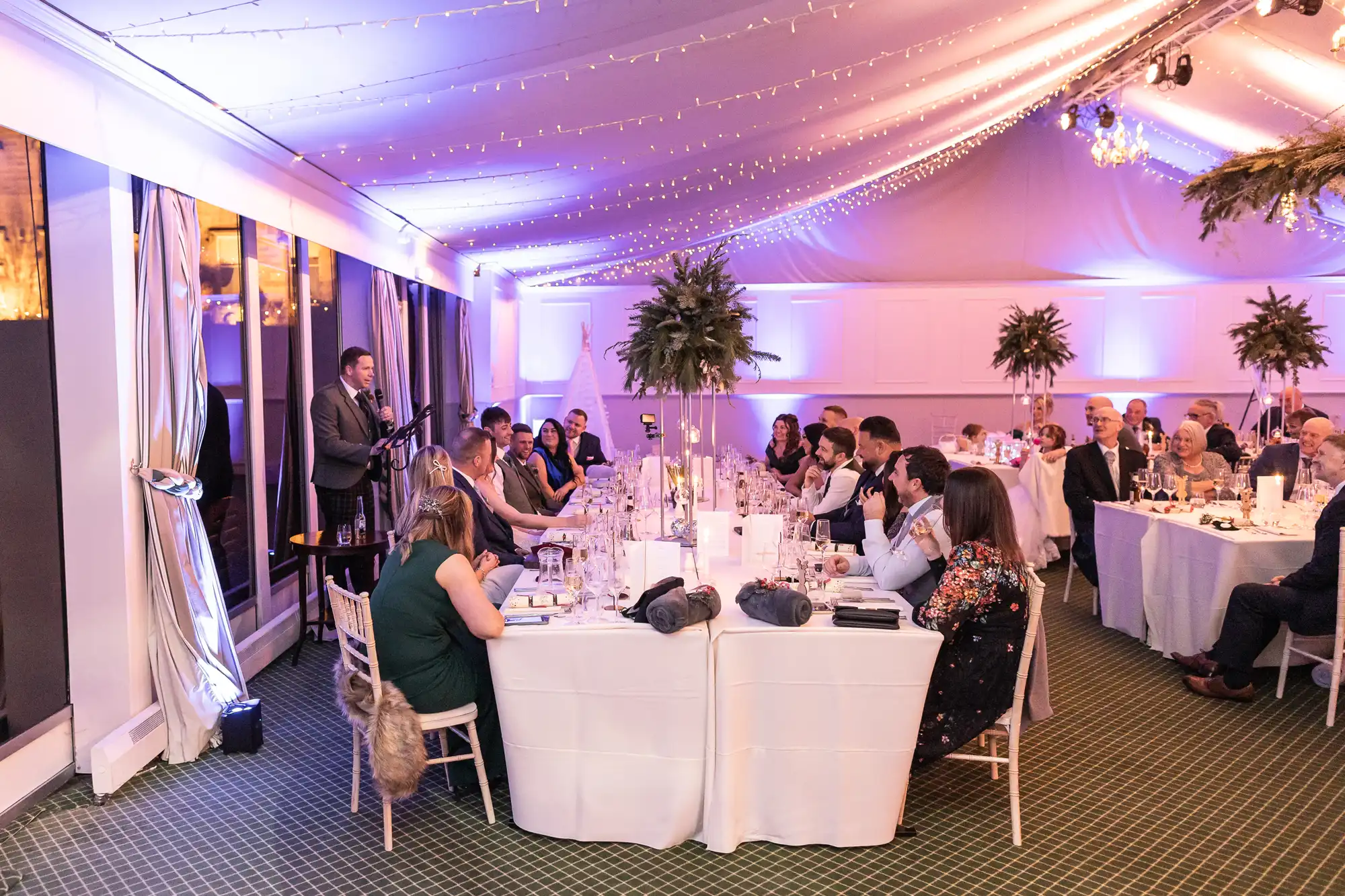 A group of people seated at decorated tables in a well-lit banquet hall, listening to a speaker who is standing near the wall. Strings of lights are draped from the ceiling.