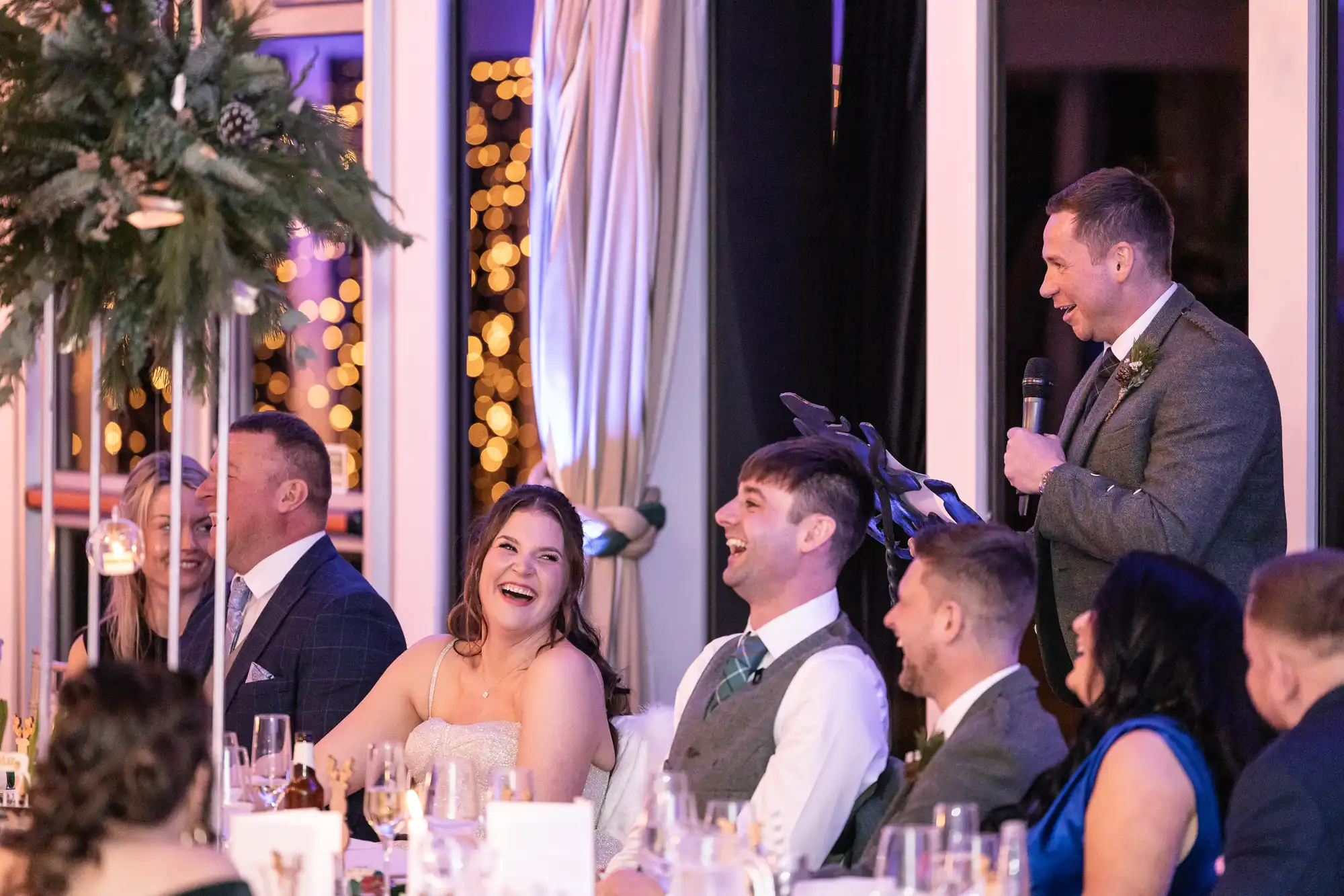 A group of people, dressed in formal attire, are seated at a banquet table, laughing and listening to a man standing and speaking into a microphone. The background is adorned with festive lights and decorations.