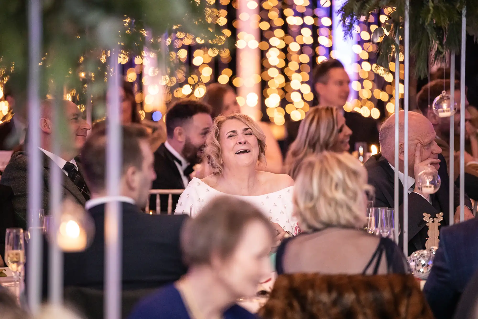 People seated at a festive event with warm string lights in the background; a woman in the center is laughing.