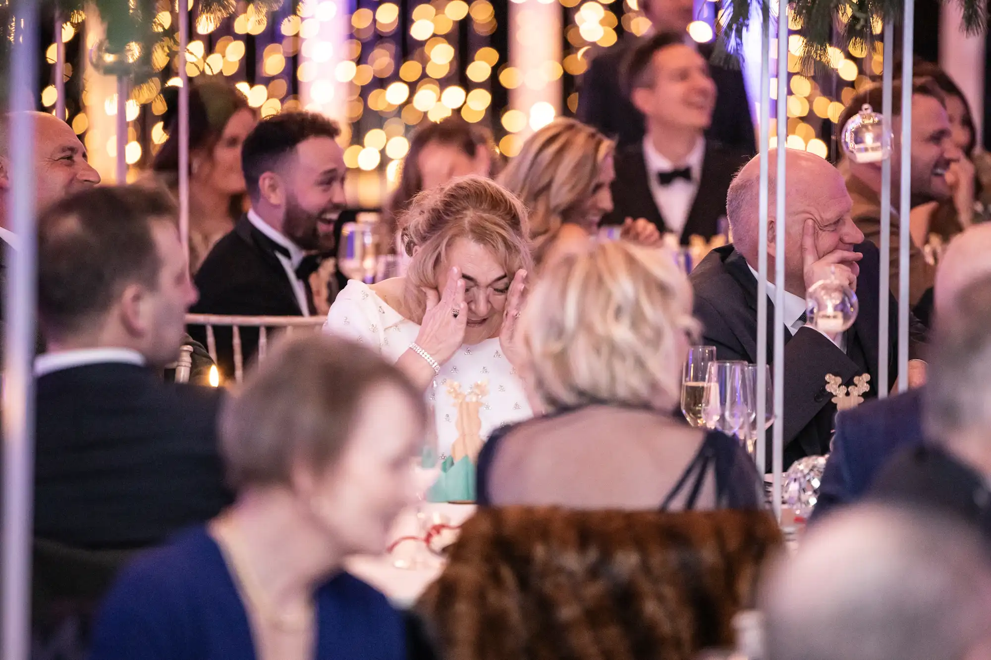 A group of people in formal attire are seated at a festive event. A woman in the center is covering her face with her hands, while others around her are smiling and looking towards the front.