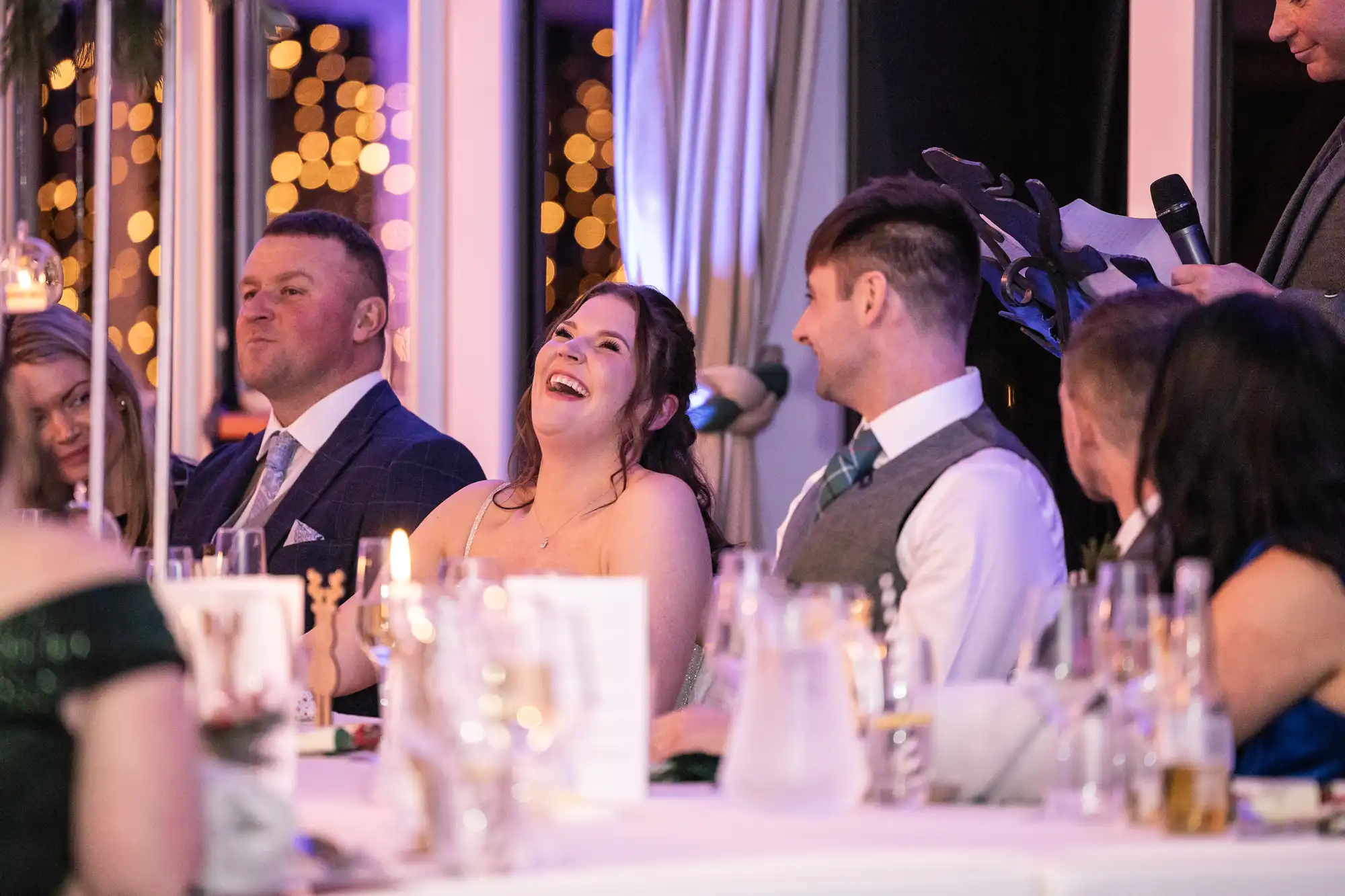 A group of people dressed formally are seated at a decorated event table, with one woman laughing. The background shows out-of-focus lights.