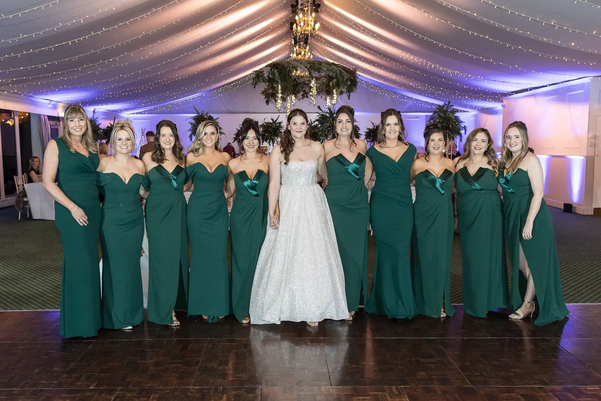 A bride in a white gown stands with ten women in matching green dresses under a tent with string lights and greenery decorations.