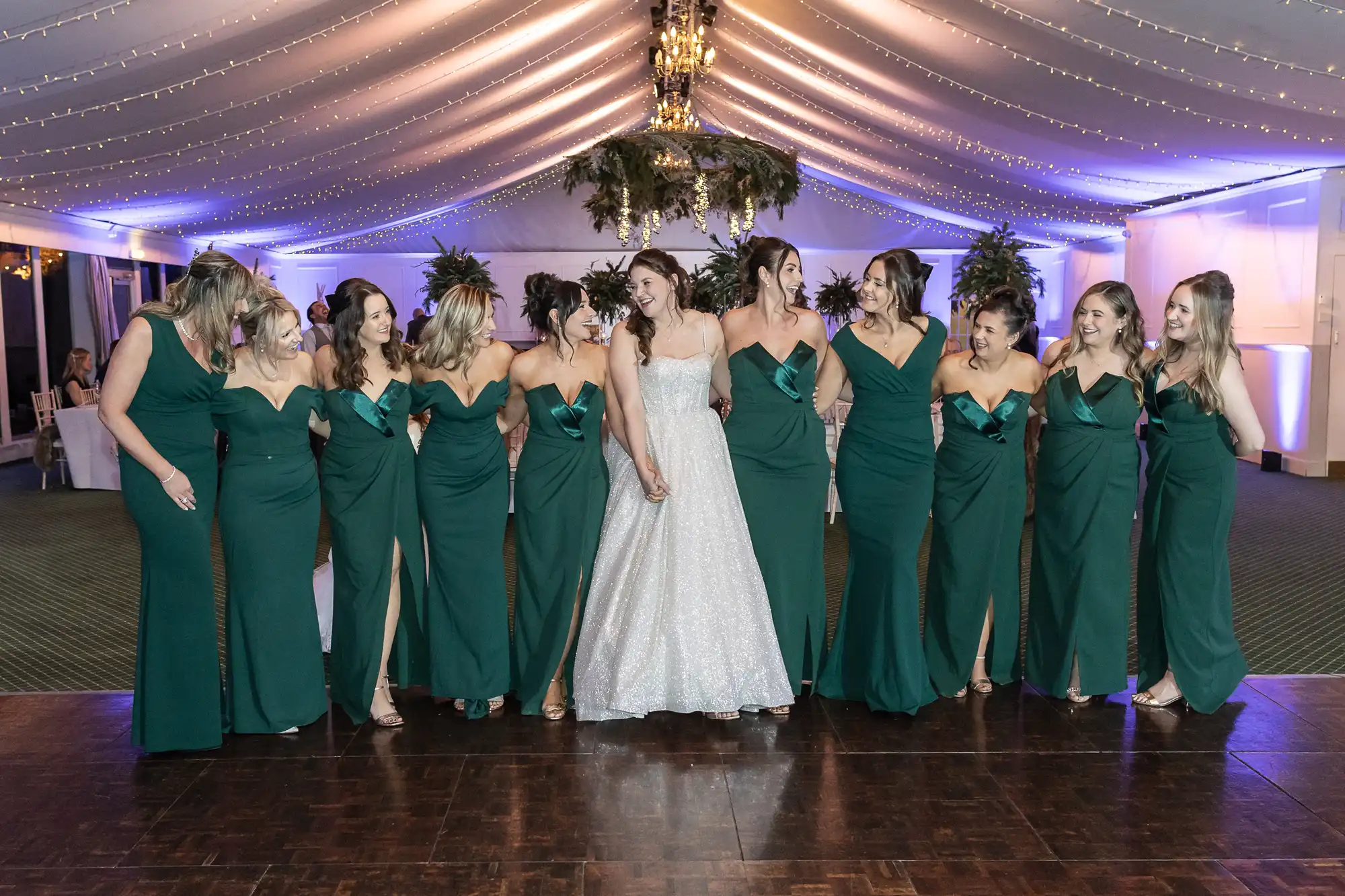 A bride in a white dress stands in the center, holding hands with bridesmaids dressed in matching green gowns under a decorated tent with a chandelier above.