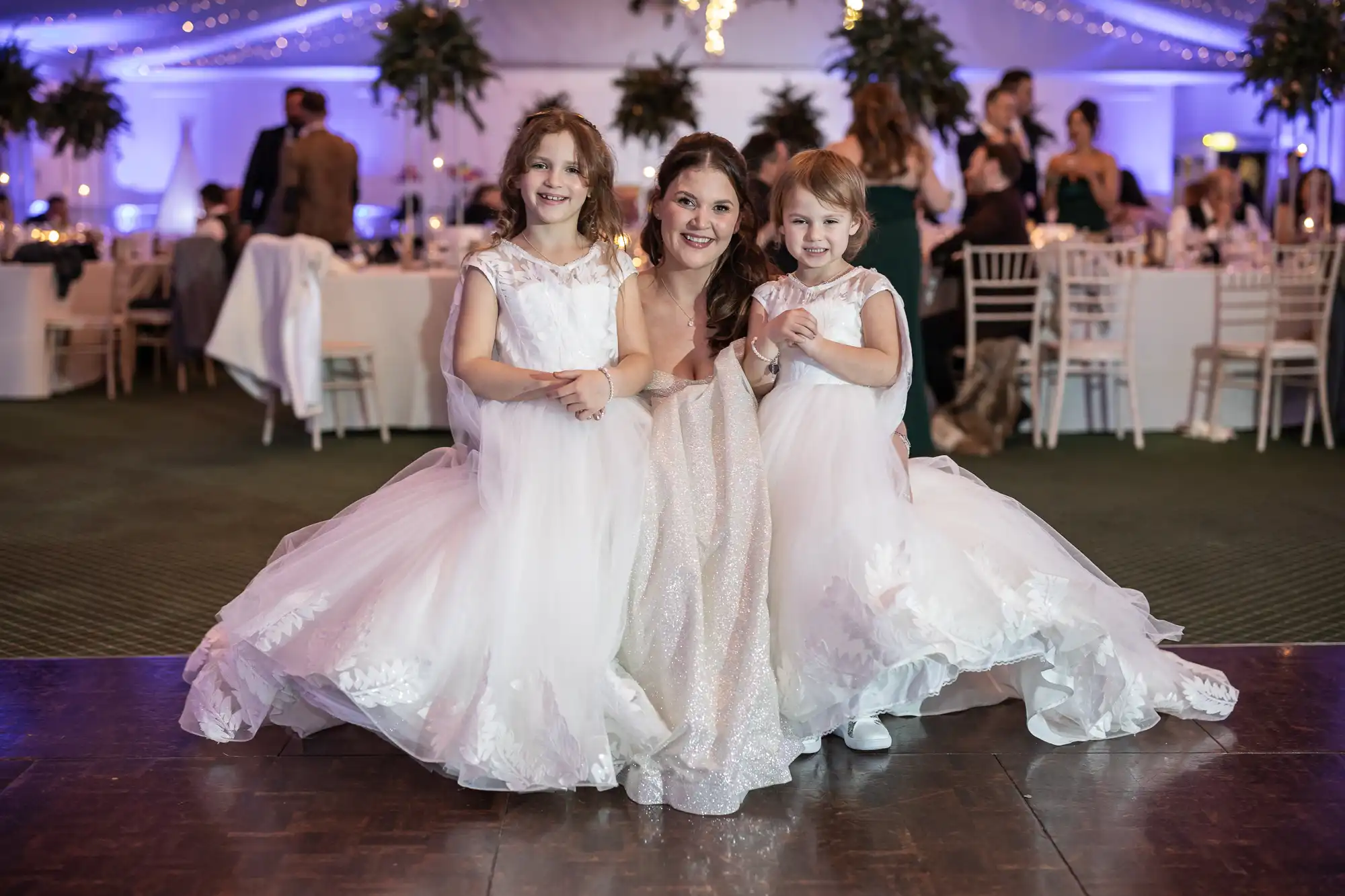 A woman in a white dress poses with two young girls, also in white dresses, at a formal event in a decorated hall with tables and guests in the background.