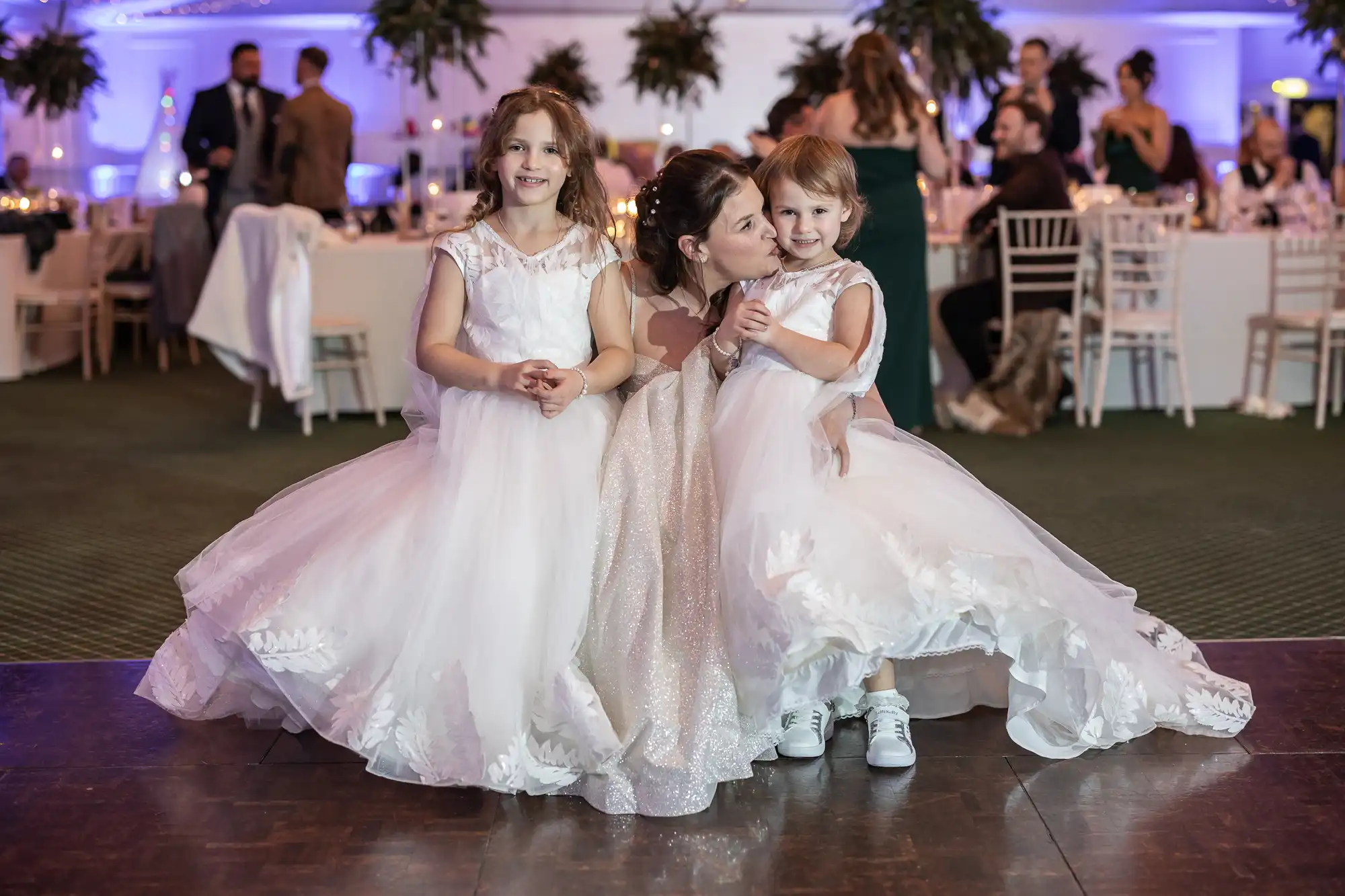 Three young girls in white dresses pose together on a dance floor at a formal event. The center girl is kissing the girl on the right, while the girl on the left smiles at the camera.