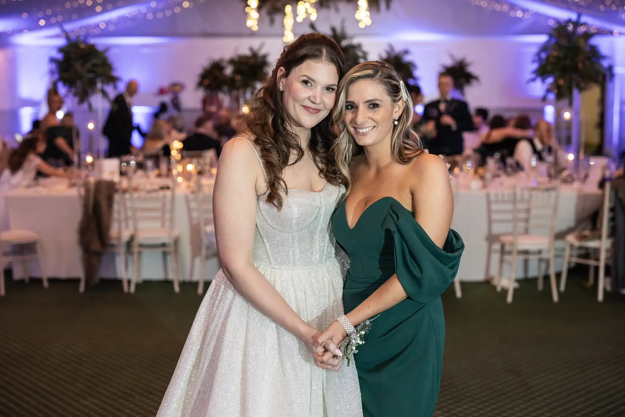 Two women dressed formally, one in a white gown and the other in a dark green dress, smiling and posing together at an indoor event with decorated tables and guests in the background.