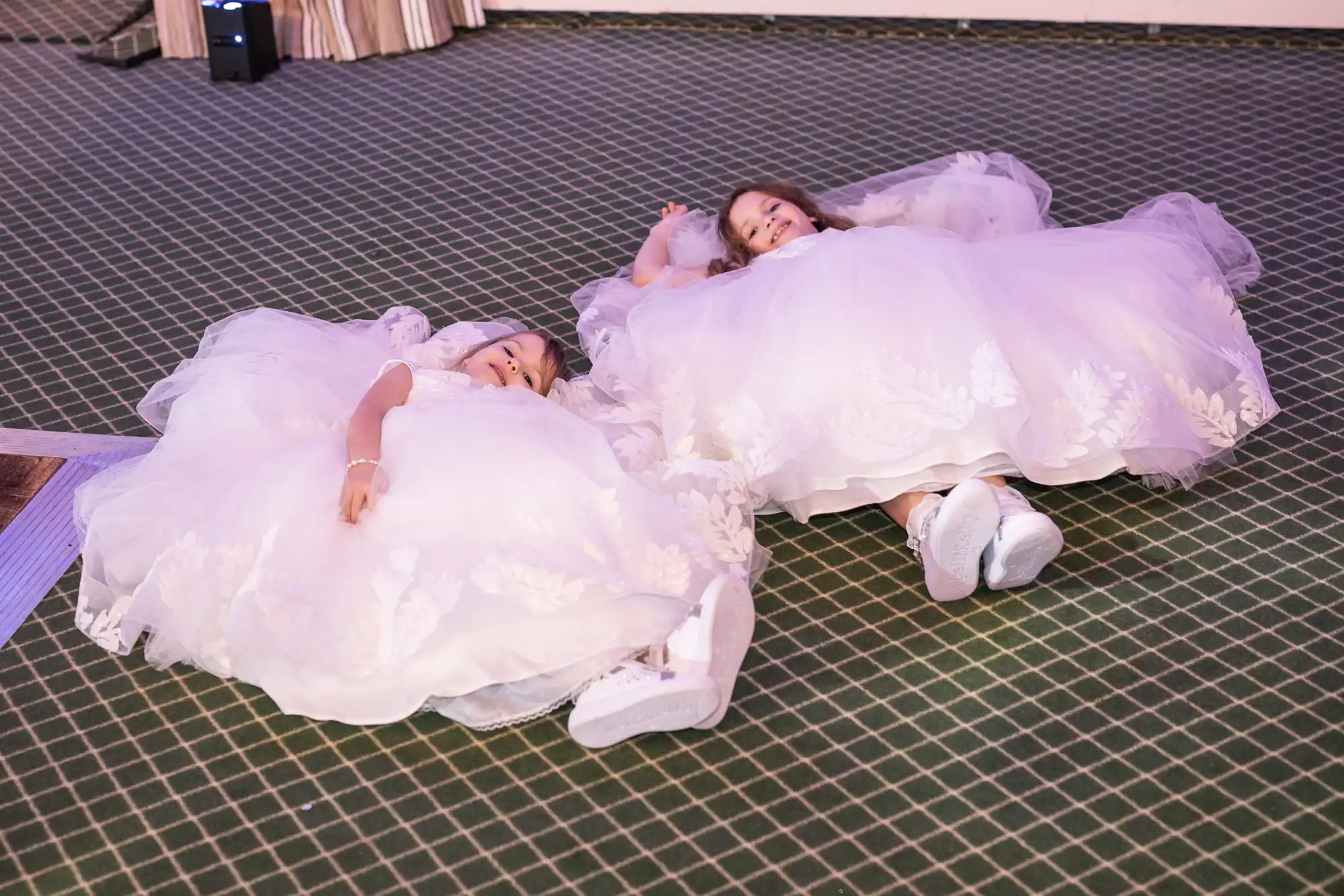 Two young children in white dresses lie on a patterned green carpet, seemingly tired or resting.