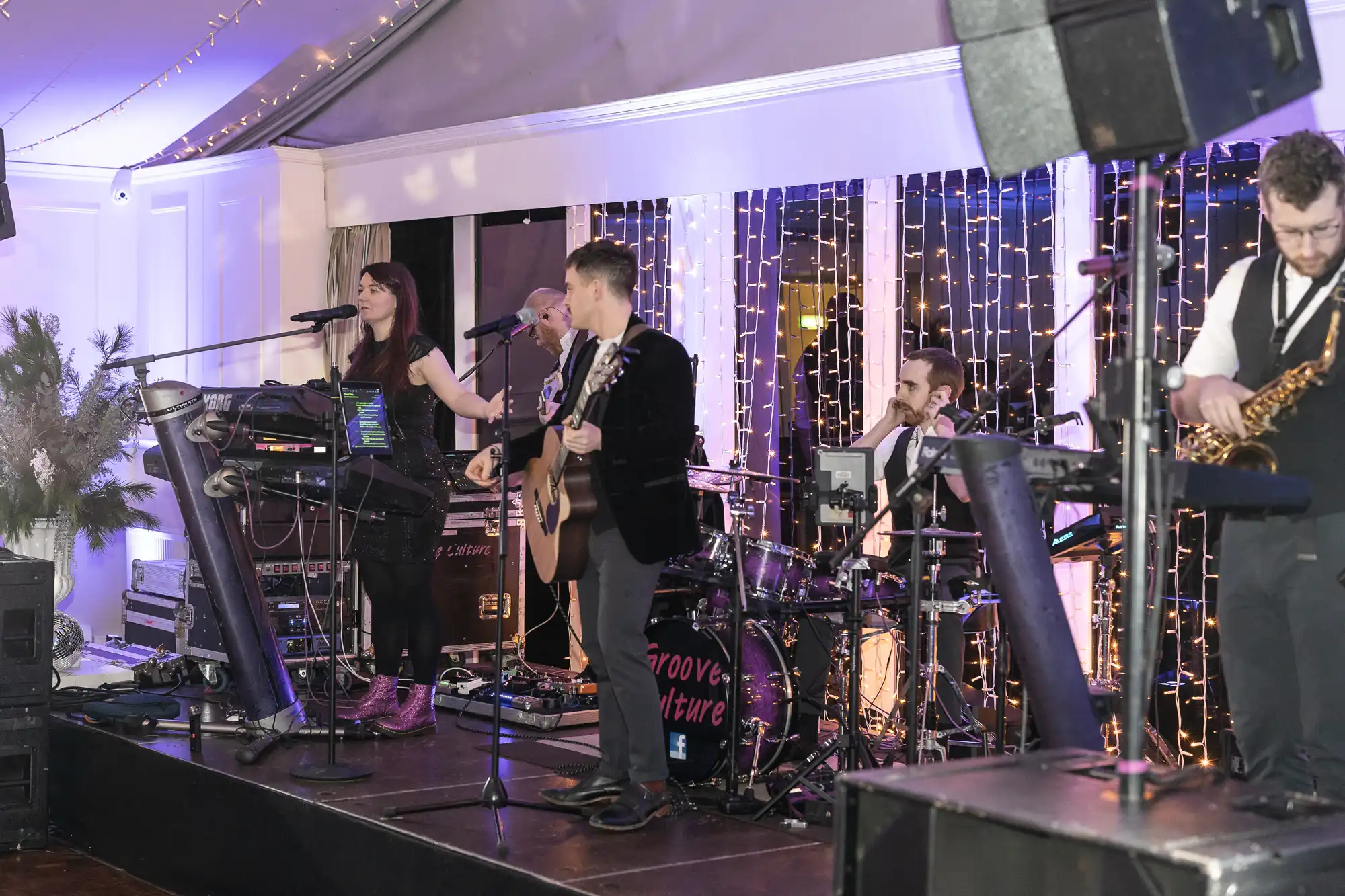 A band performs on stage with a female vocalist, guitarist, drummer, saxophonist, and keyboardist. The background features string lights and an elevated ceiling.