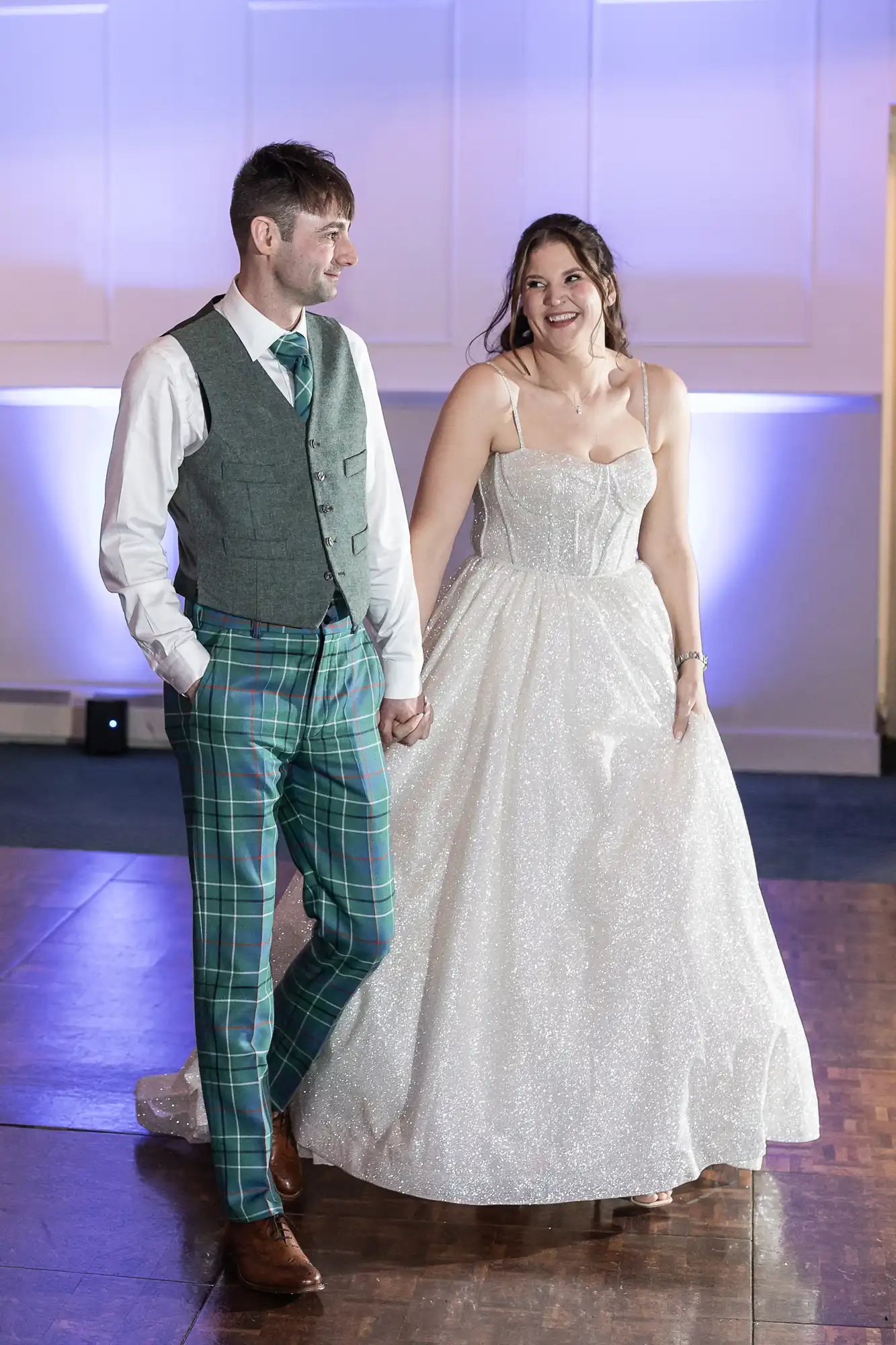 A couple holding hands while walking. The man is wearing a vest and plaid pants, and the woman is wearing a white gown.