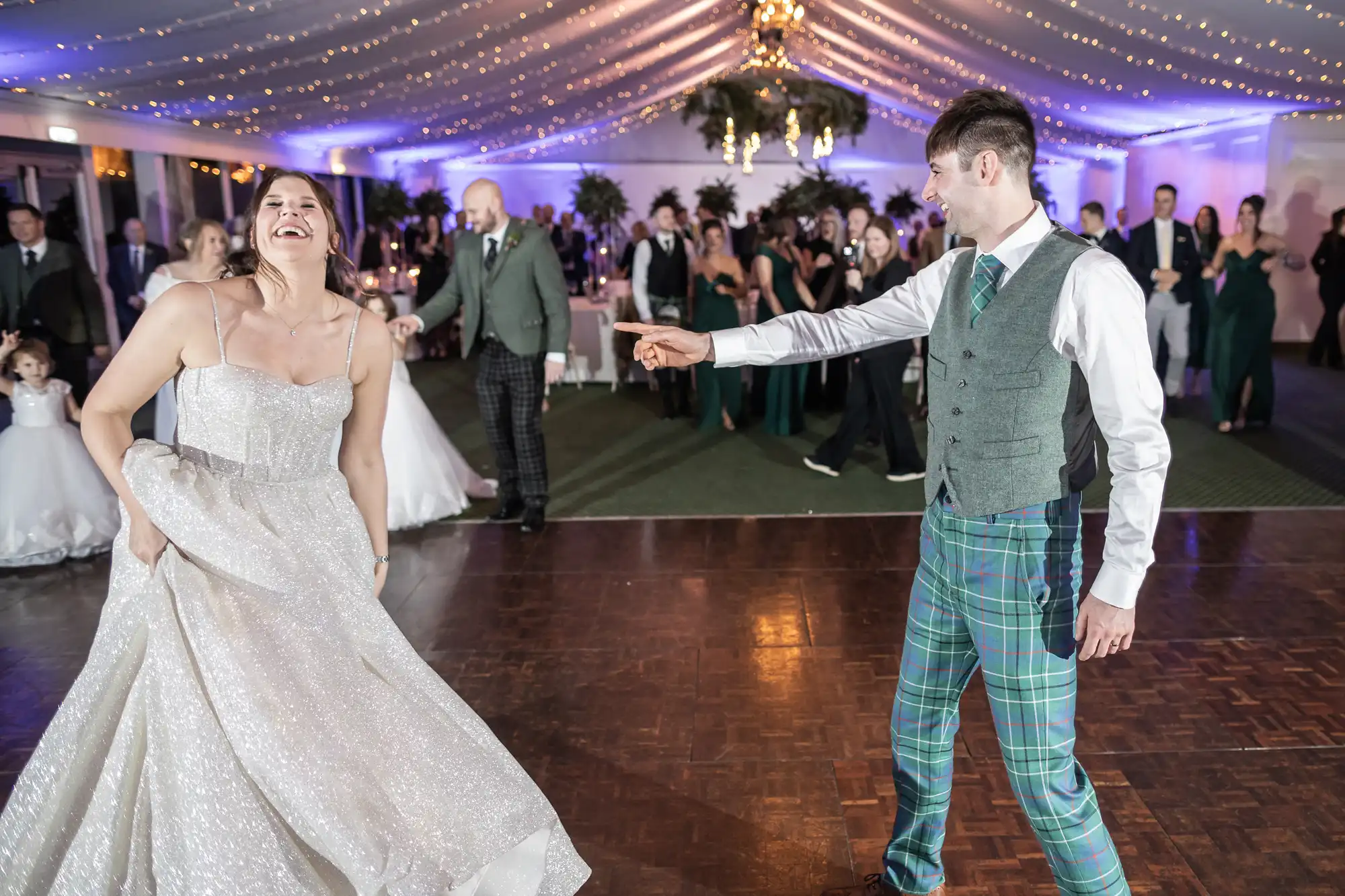 A bride and groom dance under string lights in a decorated tent as guests, including children, watch and join in the background.