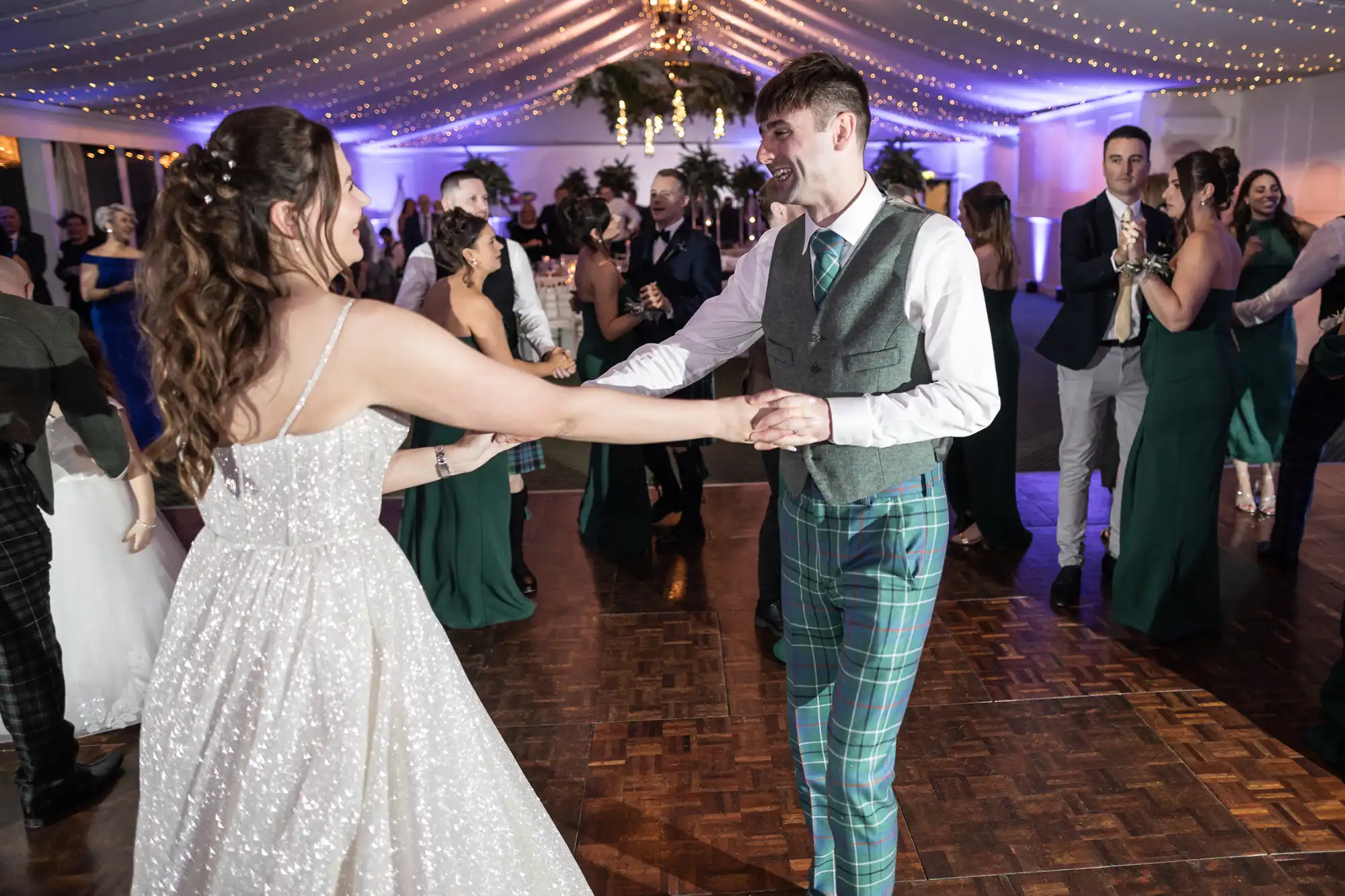 A couple dressed in formal attire dance together at a wedding reception, surrounded by other guests under a canopy adorned with string lights.