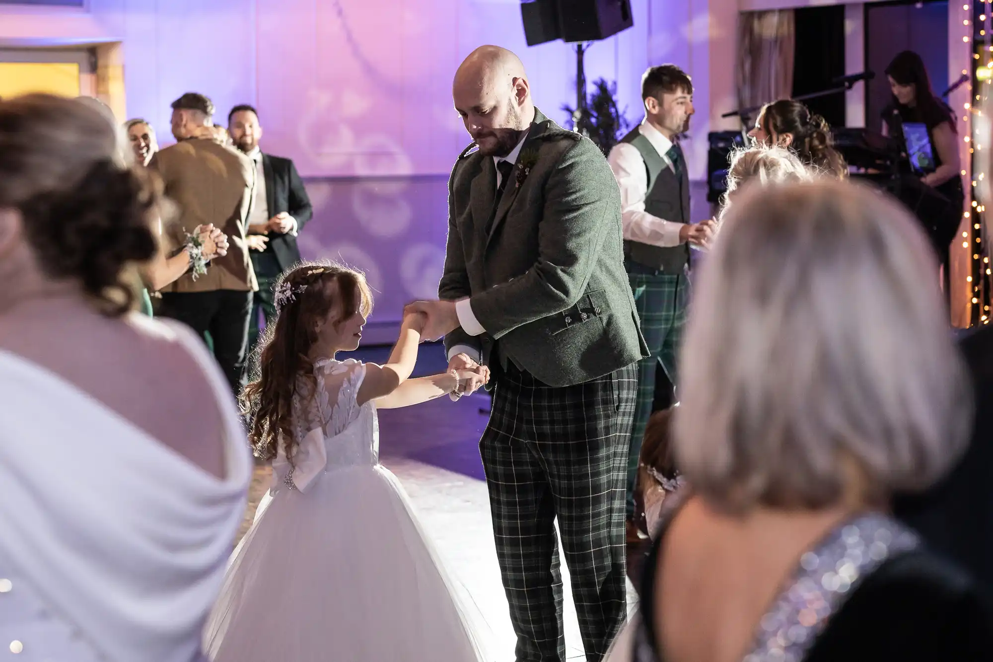 A man and a young girl in a white dress dance together in a ballroom, surrounded by other people socializing and dancing. The atmosphere is festive with soft lighting.