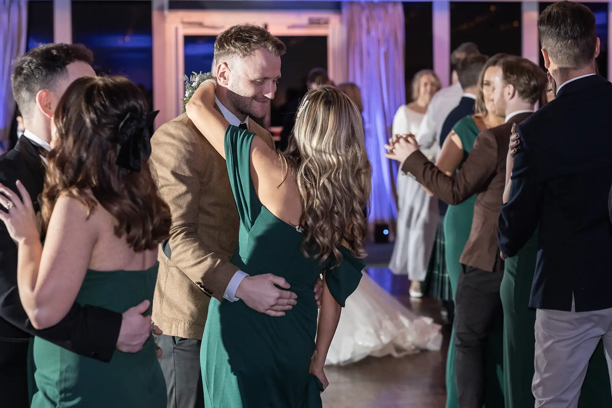 Couple dancing with others in the background at a formal event. The woman has long hair and a green dress, and the man is wearing a brown jacket.