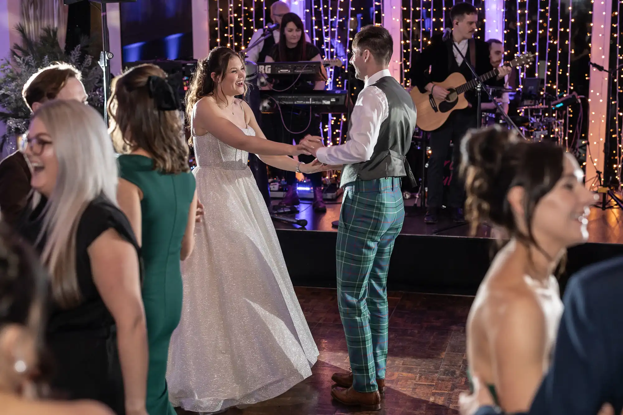 A bride and groom dance in front of a live band at a wedding reception, surrounded by guests dancing and chatting. The groom wears plaid trousers and a vest; the bride is in a white gown.