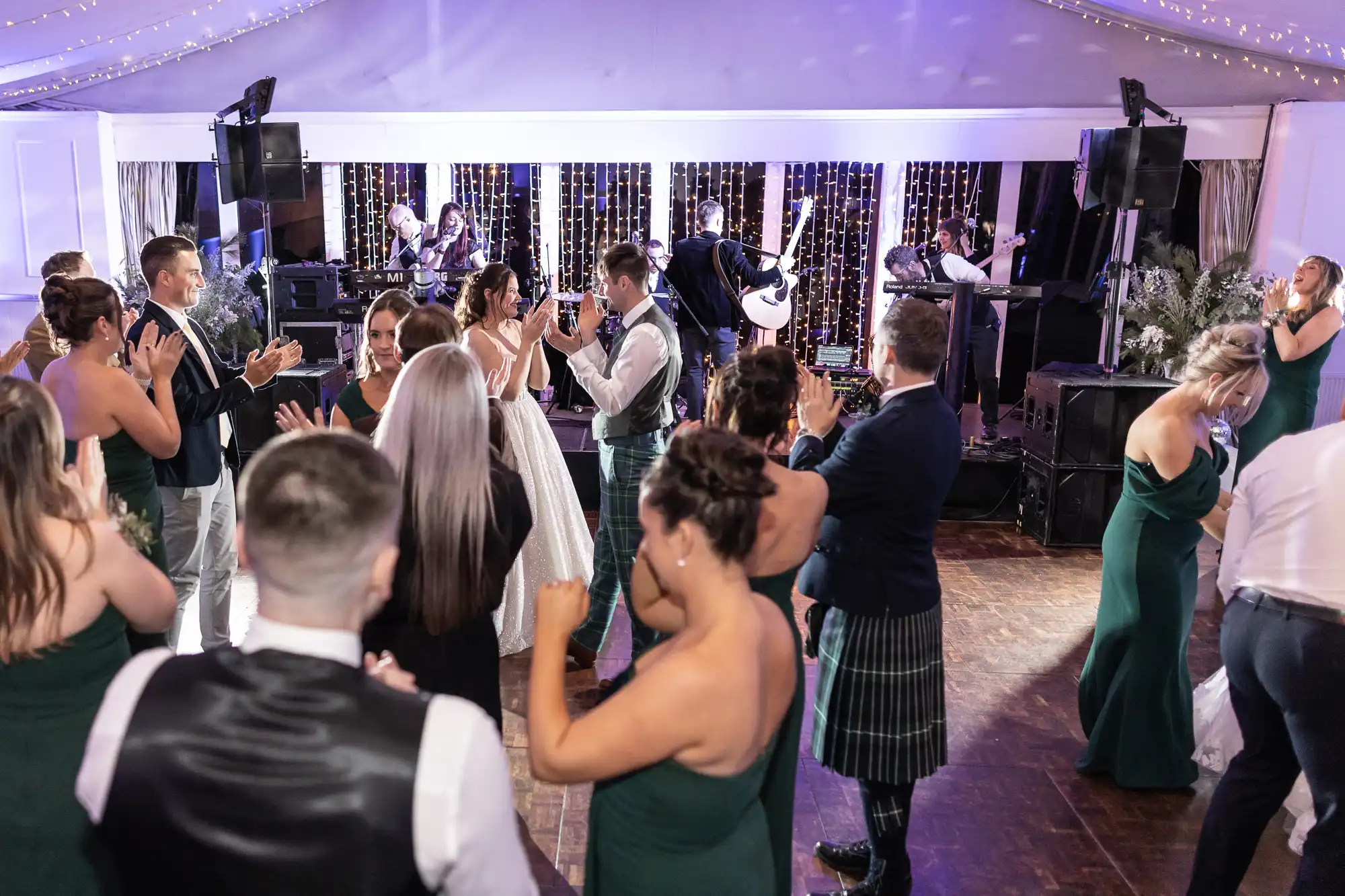 People dancing and taking photos at a lively event with a live band performing on stage in the background. The room is decorated with string lights.