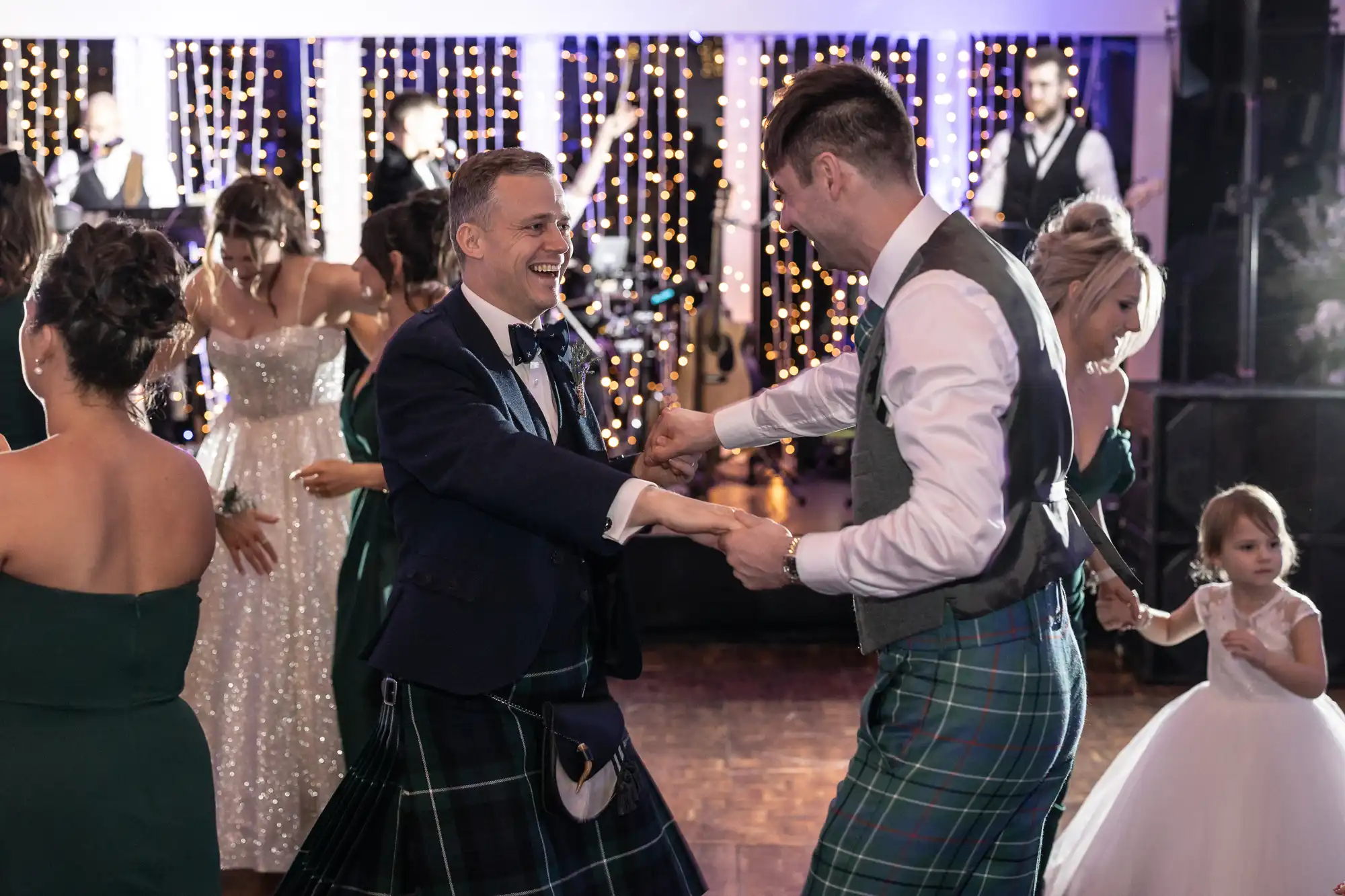 Two men in kilts dance at a wedding reception. One is wearing a blue jacket with a bow tie and the other a vest and tie. Other guests, including two women in dresses and a child, are also dancing.