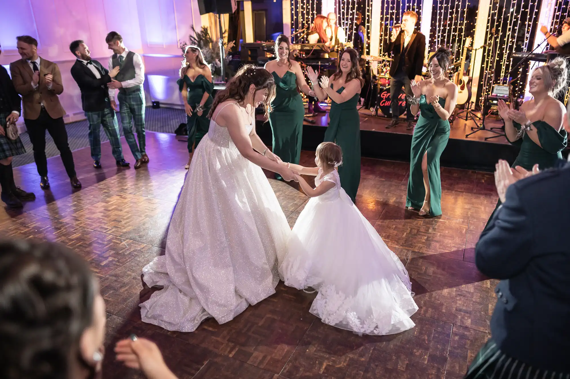 A bride in a white dress dances with a young girl also in a white dress, surrounded by guests clapping and smiling at an indoor event with a live band in the background.