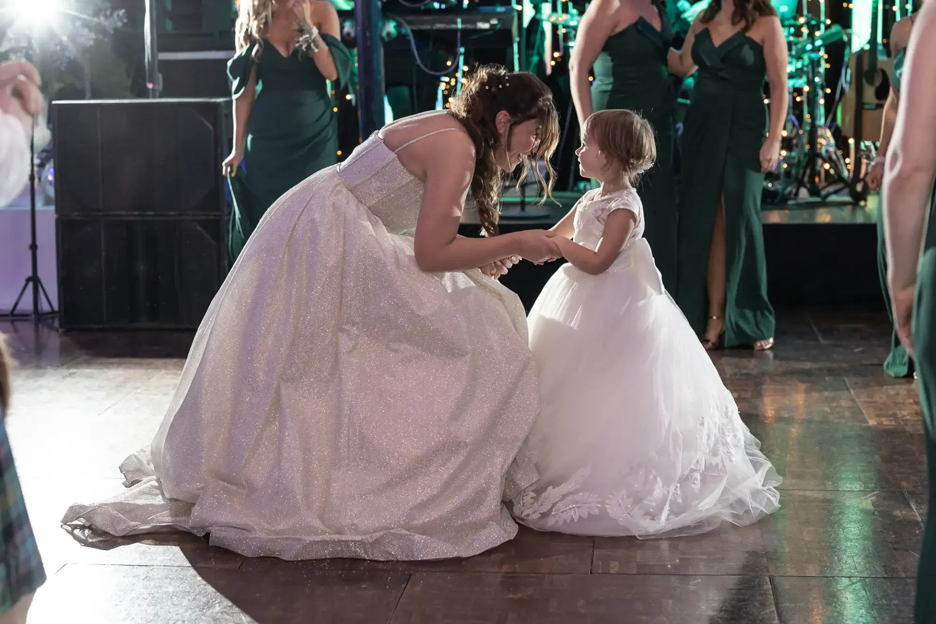 A woman in a white gown bends down to hold hands with a young girl in a white dress on a dance floor, surrounded by people in green dresses. A band is performing in the background.