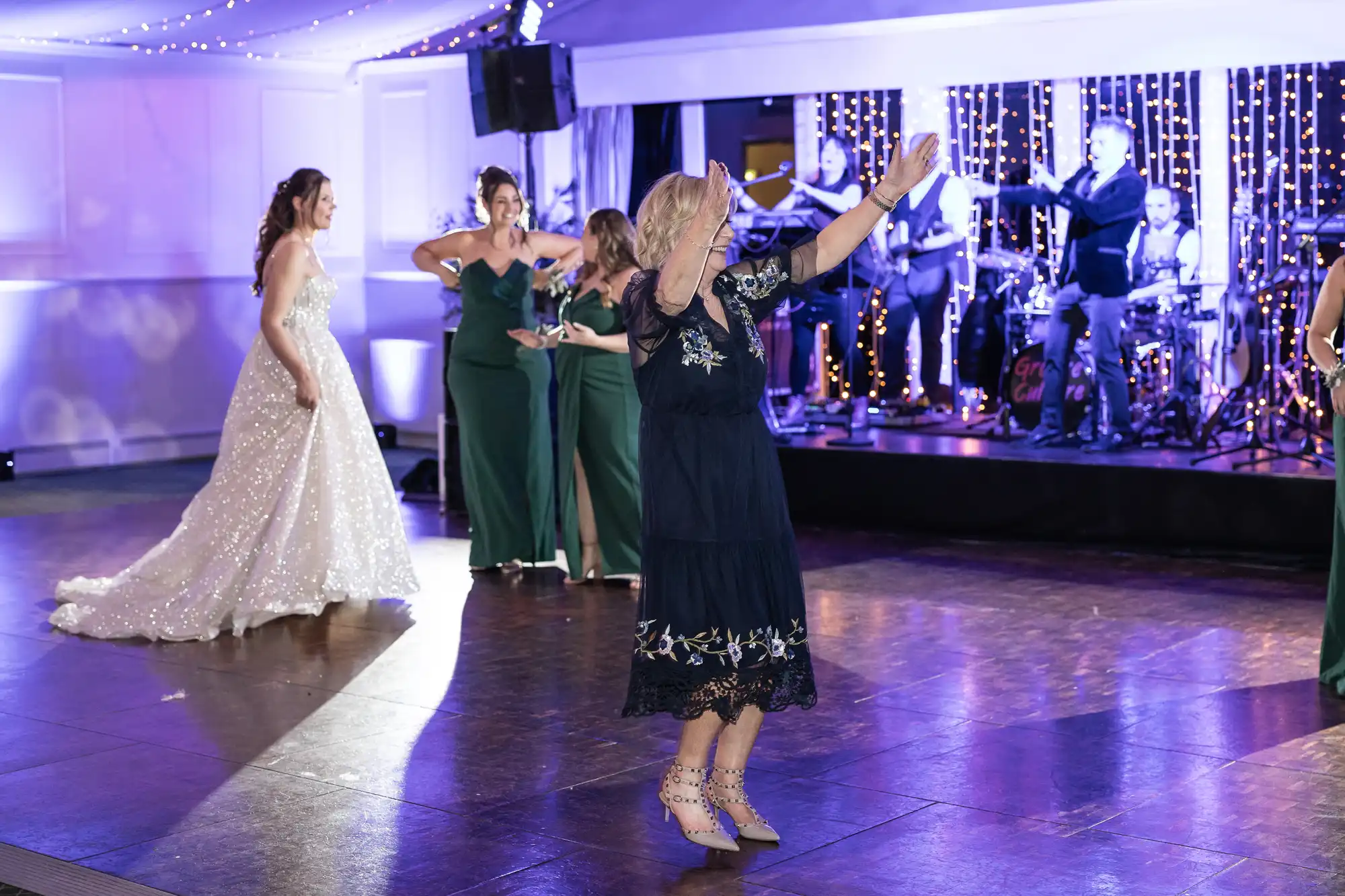 Guests are dancing on a wooden floor at an event while a live band performs on stage in the background. A bride in a white gown is visible on the left.