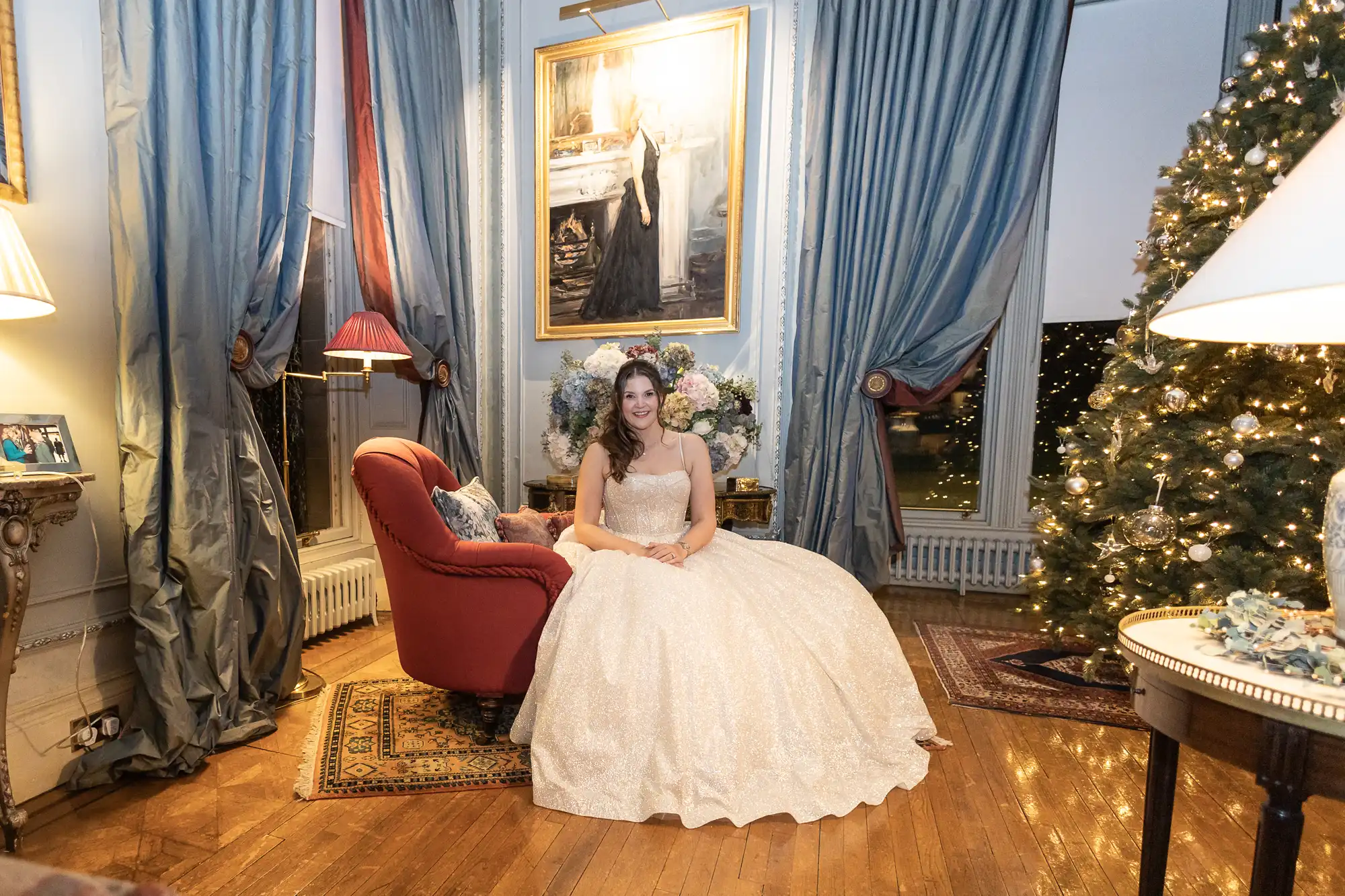 A woman in a white gown sits on a red chair in an elegant room with blue curtains, a decorated Christmas tree, and a large portrait on the wall.