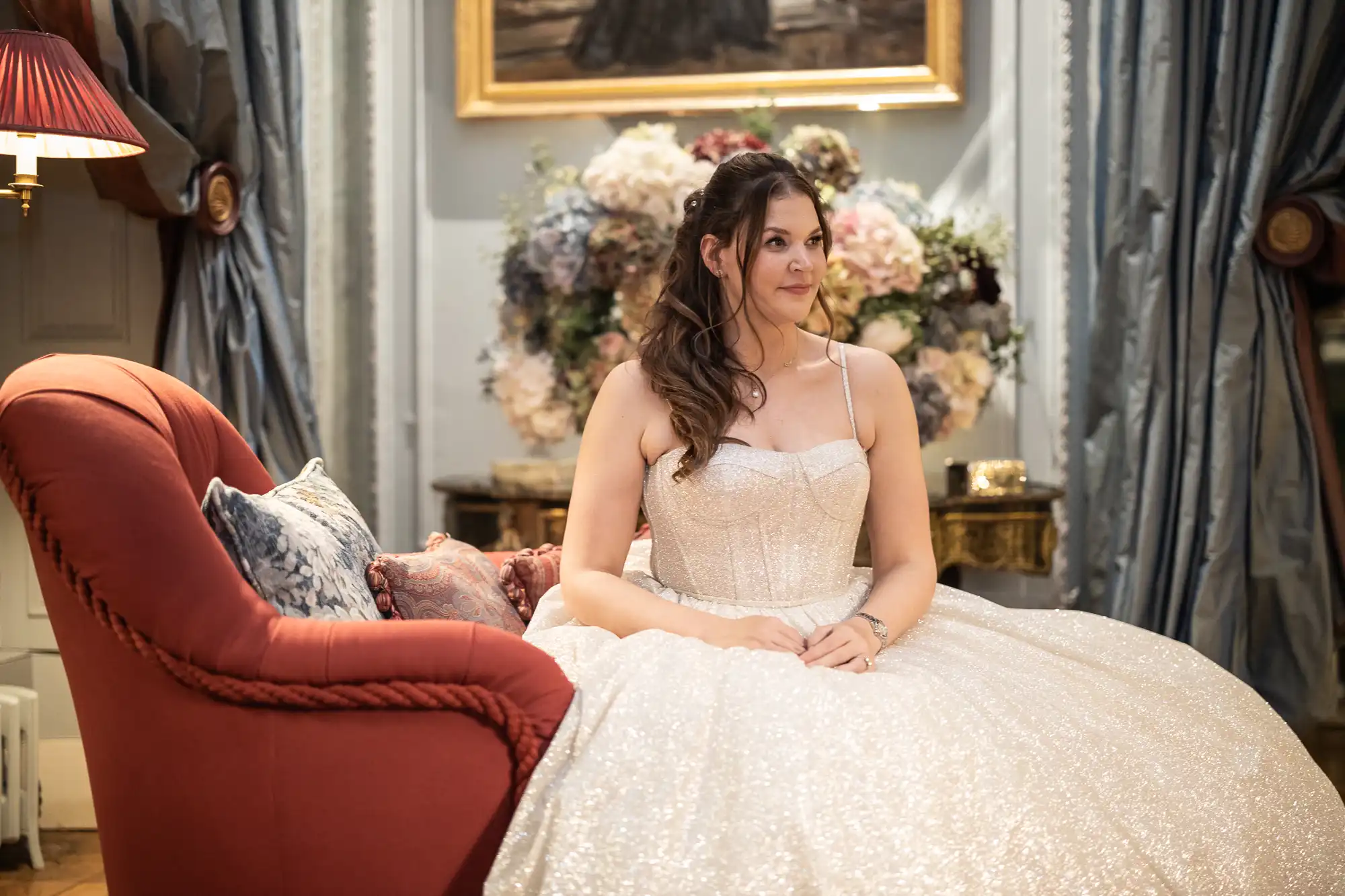 A woman in a white sparkling dress sits on a red couch in an elegantly decorated room, with flowers and a painting in the background.