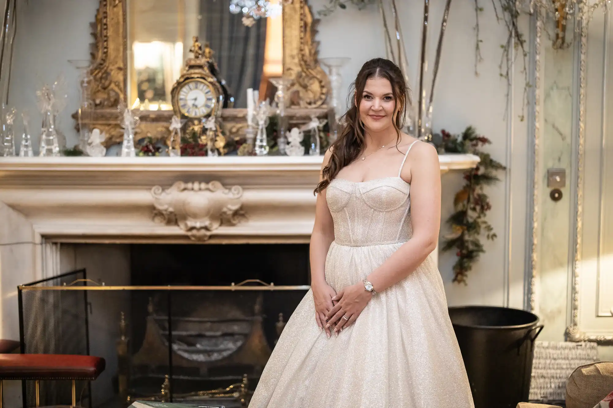 Woman in a white, strapless gown stands in front of an ornate fireplace, decorated with a clock and other decorative items.