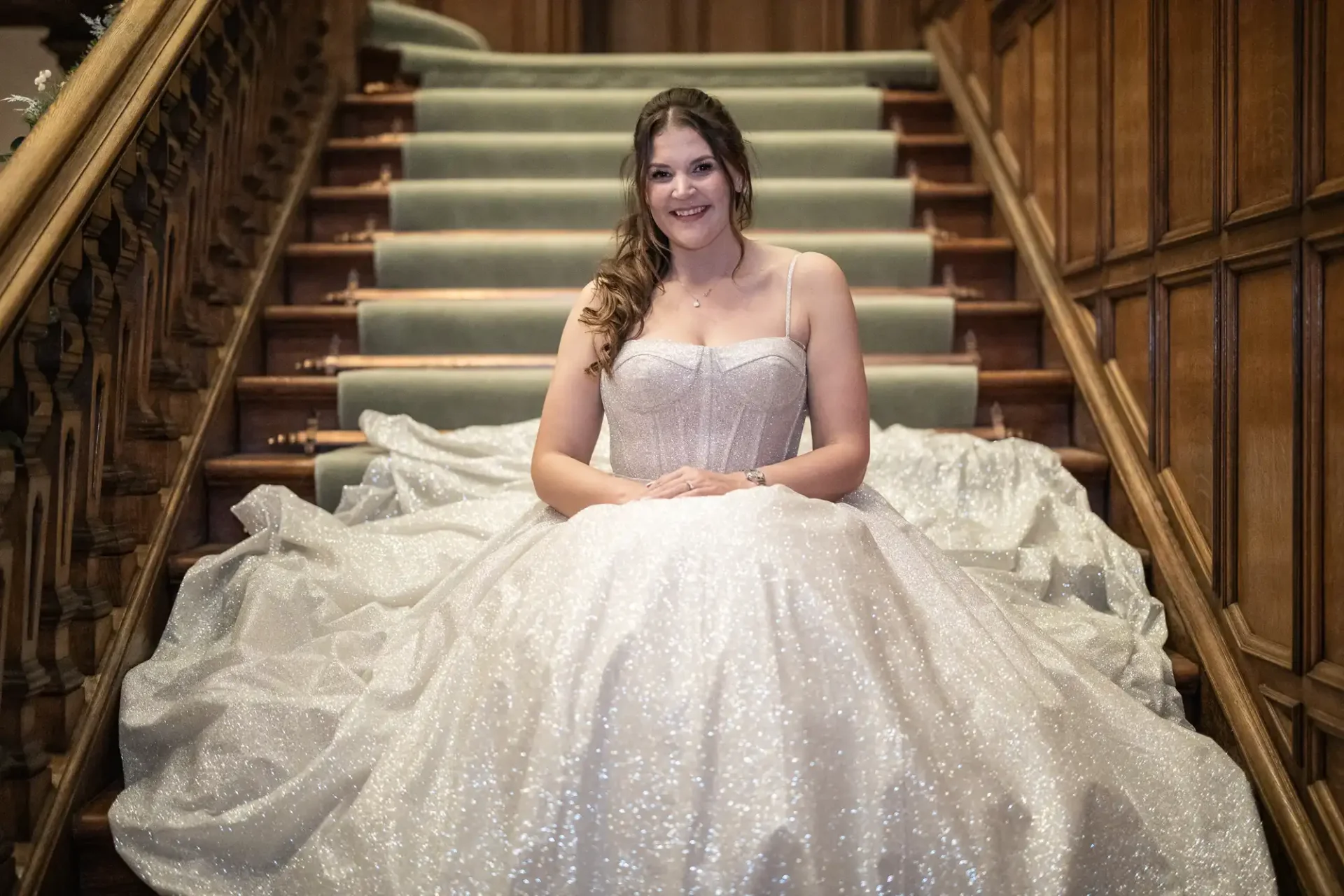 A woman in a sparkling white gown sits on a staircase with wooden paneling in the background, smiling at the camera. .
