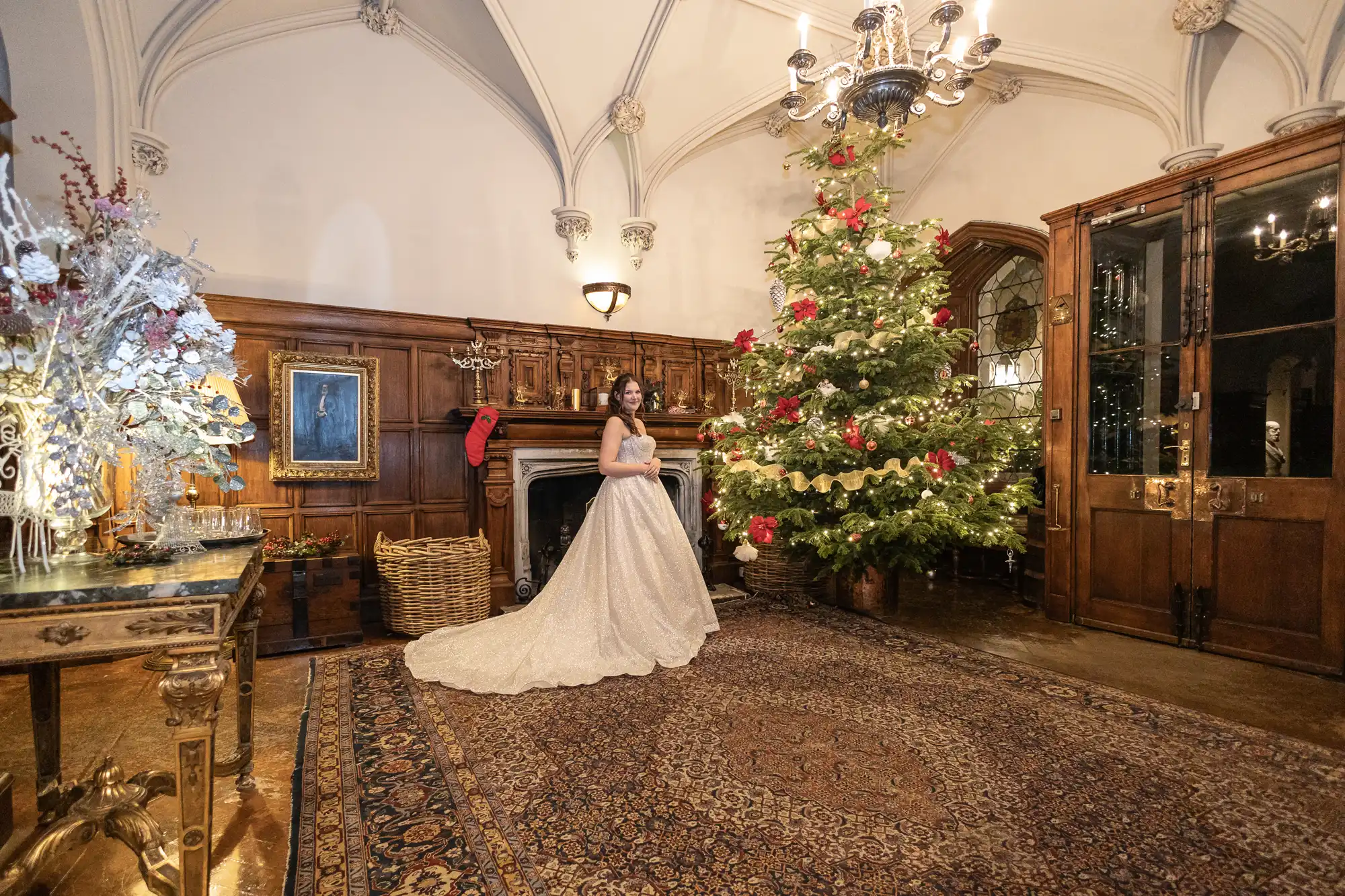 A woman in a white gown stands beside a decorated Christmas tree in a large, ornate room with wooden panels and a chandelier.