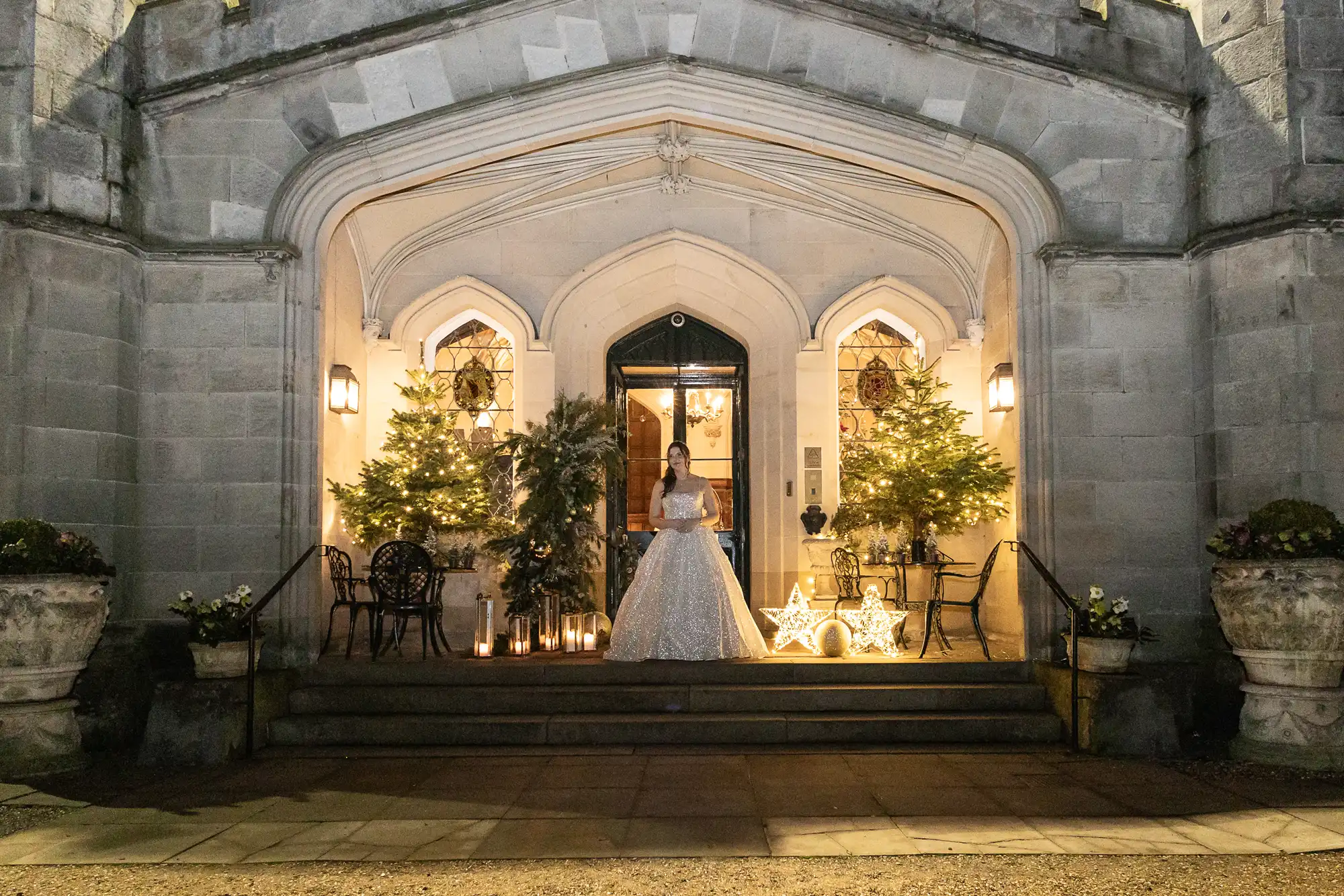 A person in a white, intricately detailed gown stands at the entrance of a grand, Gothic-style building, flanked by two Christmas trees and lit by warm lighting and decorative elements.