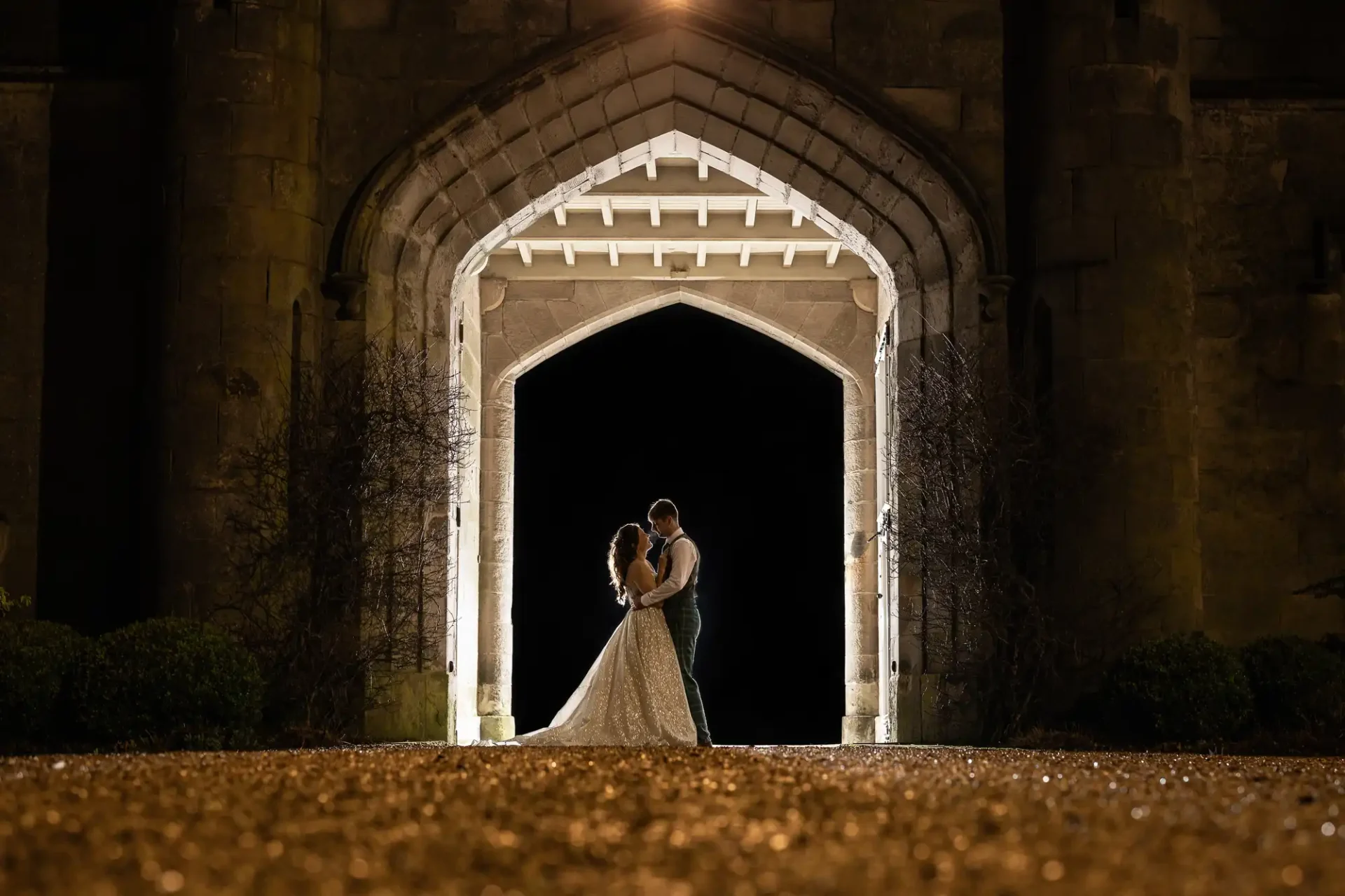 A couple stands embraced under a gothic archway at night, illuminated by lights behind them, creating a silhouette against the dark background.