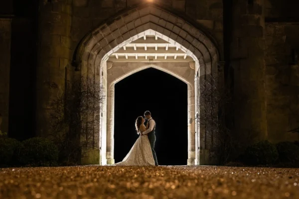 Dundas Castle wedding photography - capturing real moments at historic Scottish castles. A couple stands embraced under a gothic archway at night, illuminated by lights behind them, creating a silhouette against the dark background.