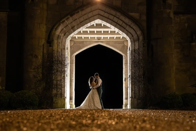 Dundas Castle Wedding: A couple stands embraced under a gothic archway at night, illuminated by lights behind them, creating a silhouette against the dark background.