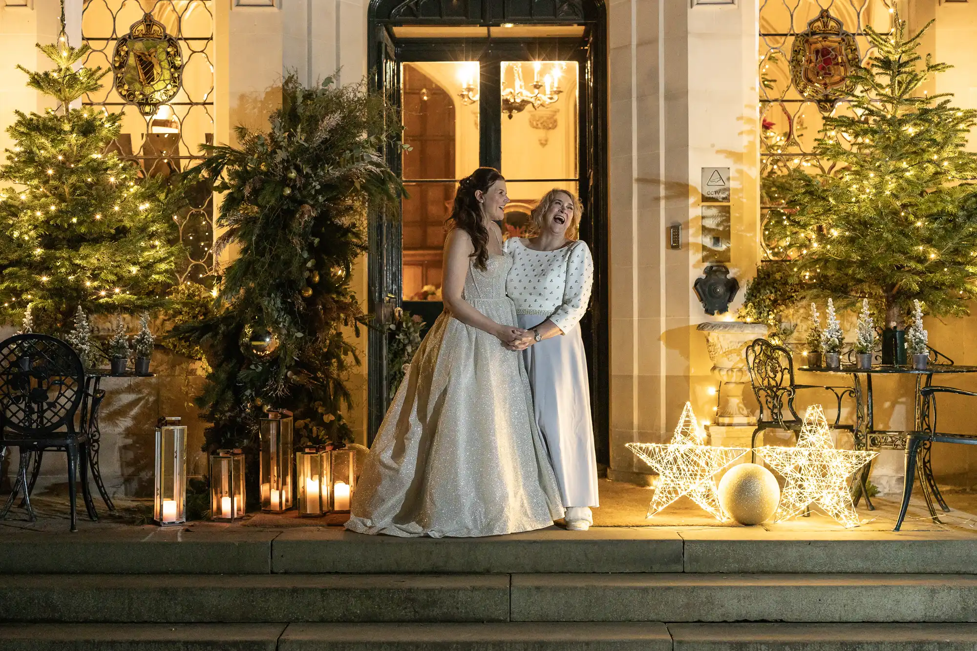 Two women in elegant gowns stand holding hands and smiling by the illuminated entryway of a building adorned with Christmas trees and decorations.