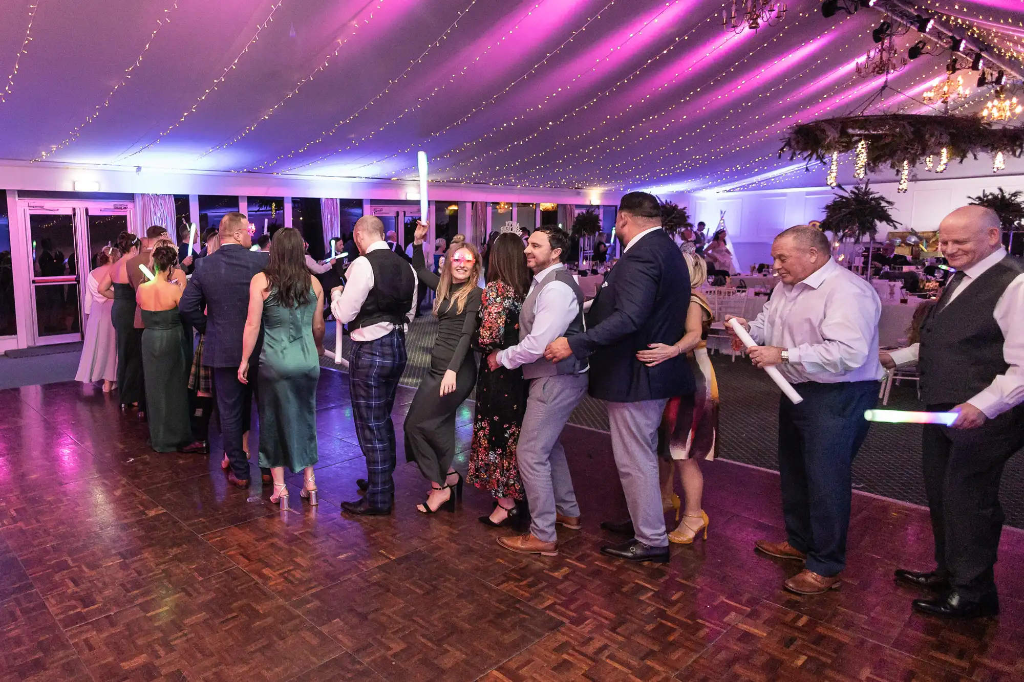 A group of people in formal attire are dancing in a conga line under a tent with string lights and pink and blue lighting.