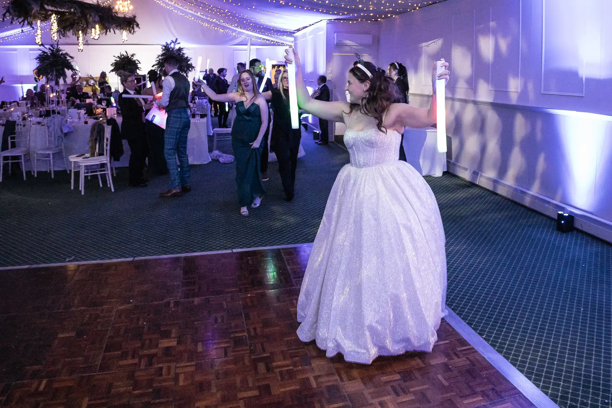 A bride in a white dress dances on an empty dance floor while guests mingle and dance in the background at a wedding reception.