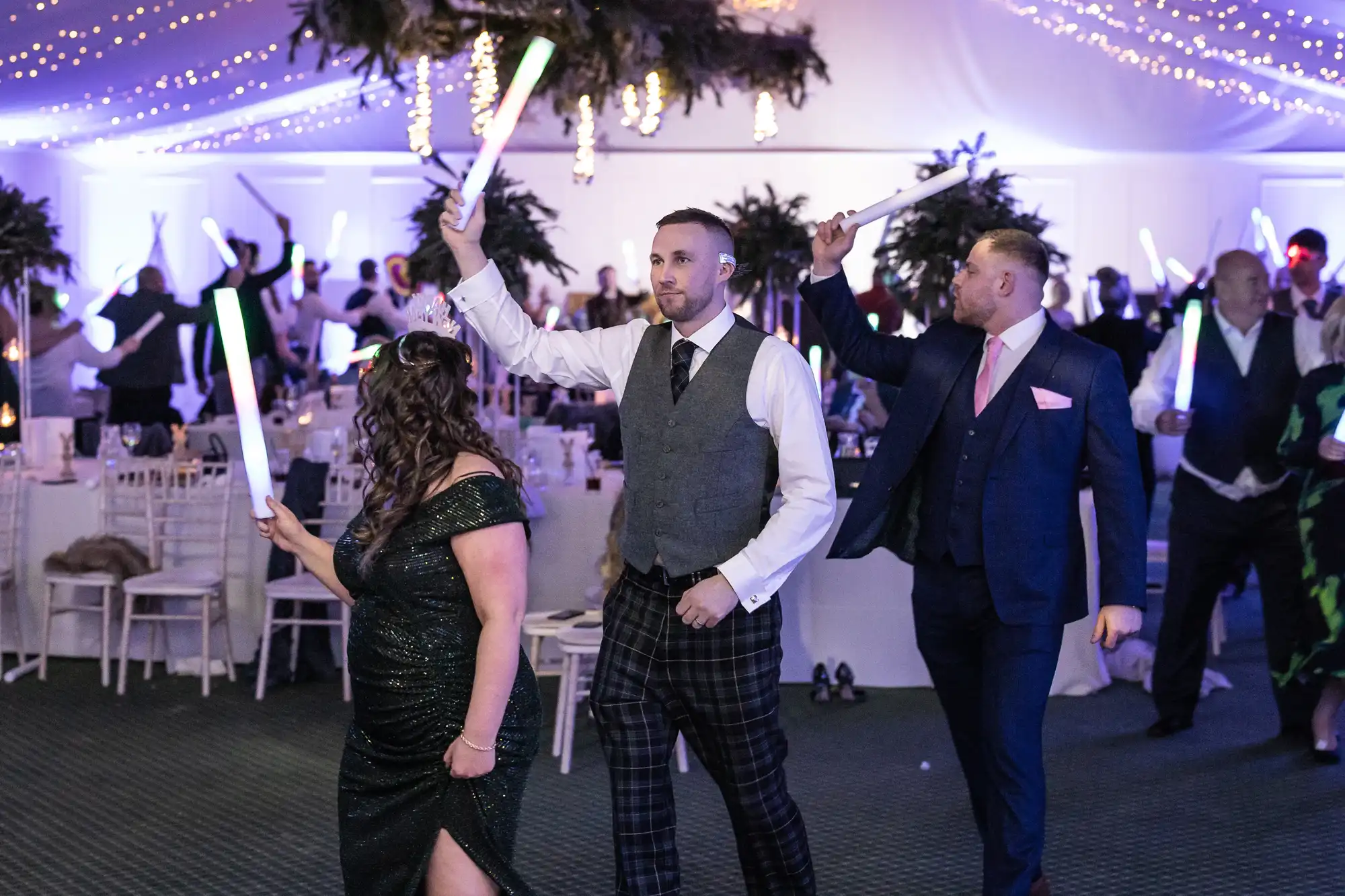 People in formal attire are walking while holding light sticks at an indoor event with tables and decorative lighting.