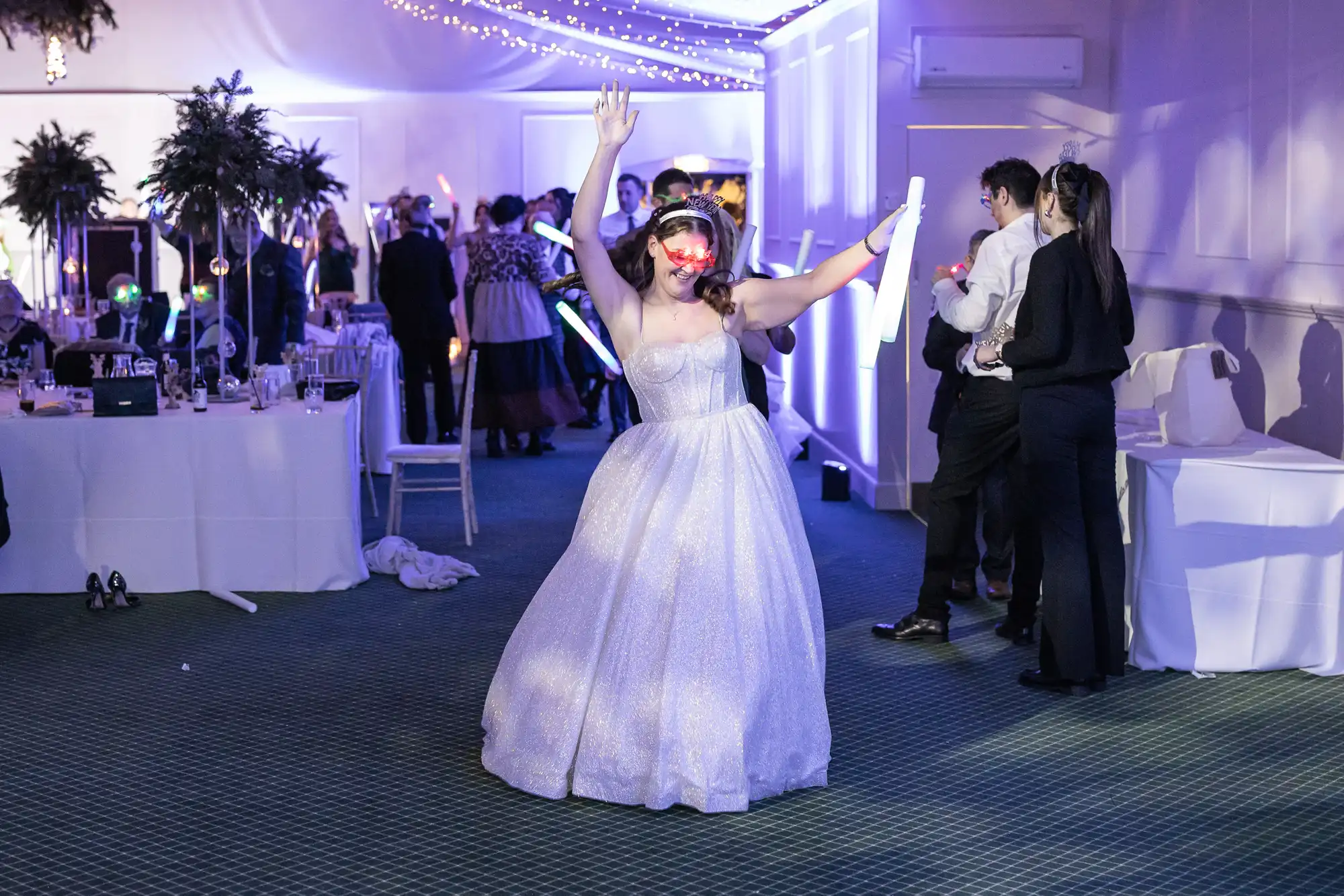 A person in a sparkling dress dances enthusiastically in a dimly lit room with party guests and decor in the background.