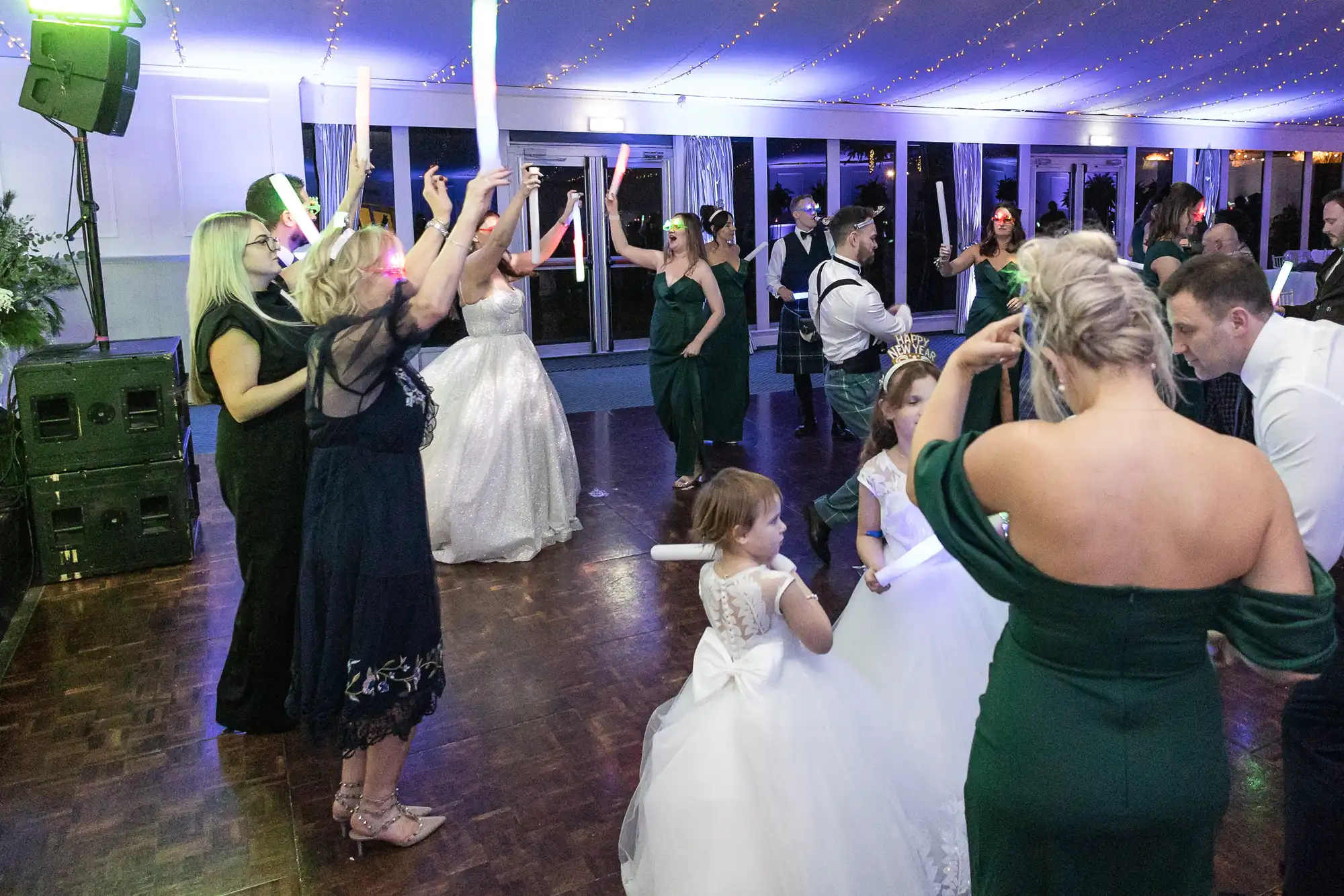 People, including a bride and two flower girls, dancing and holding glow sticks at a wedding reception.