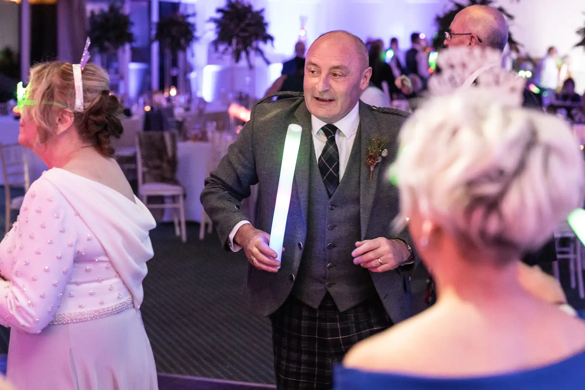 A man in a grey suit holds a glowing stick while dancing at a festive event, surrounded by people wearing light-up accessories.