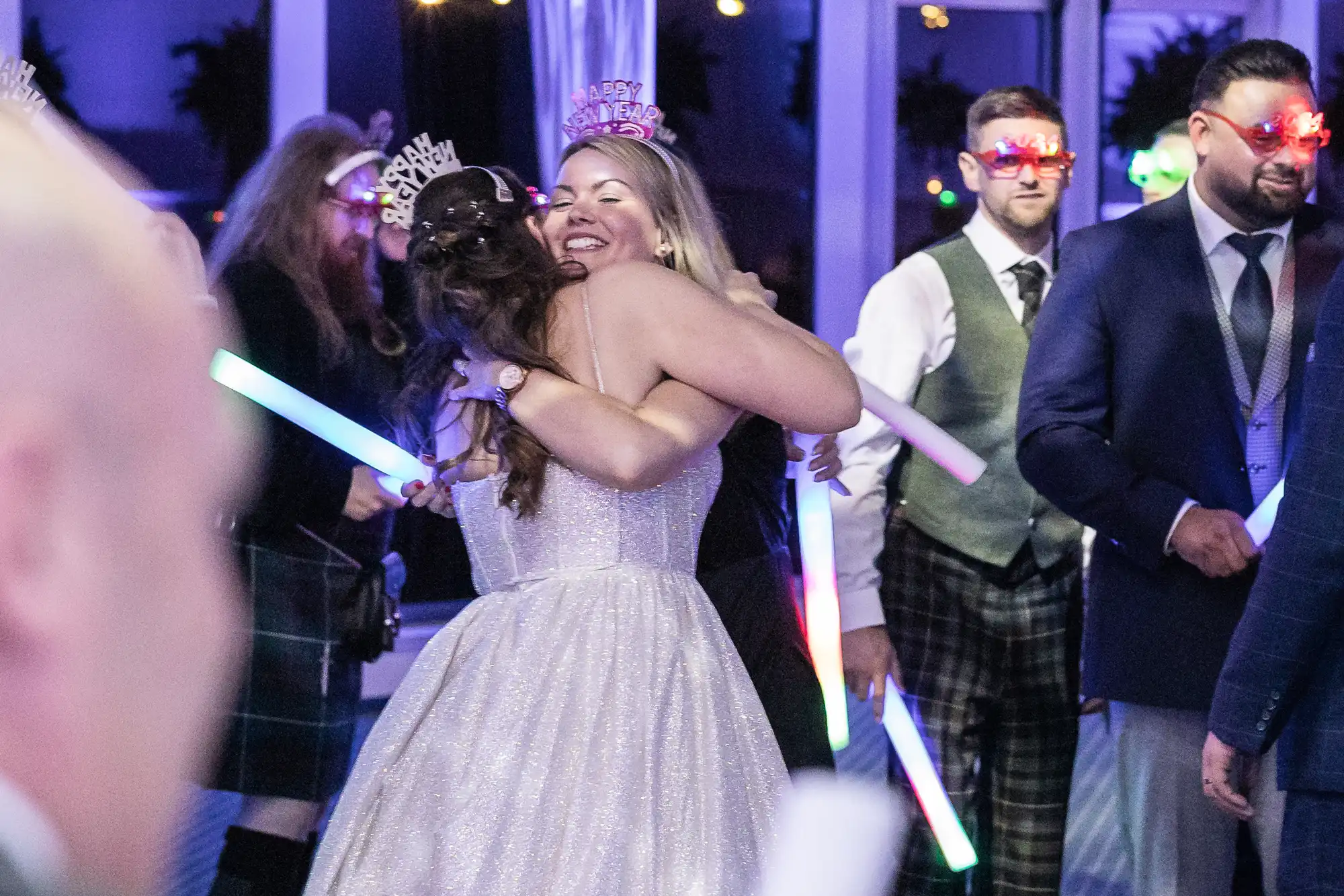 People celebrating New Year's Eve at a party, with two women hugging and others holding light sticks and wearing festive accessories.