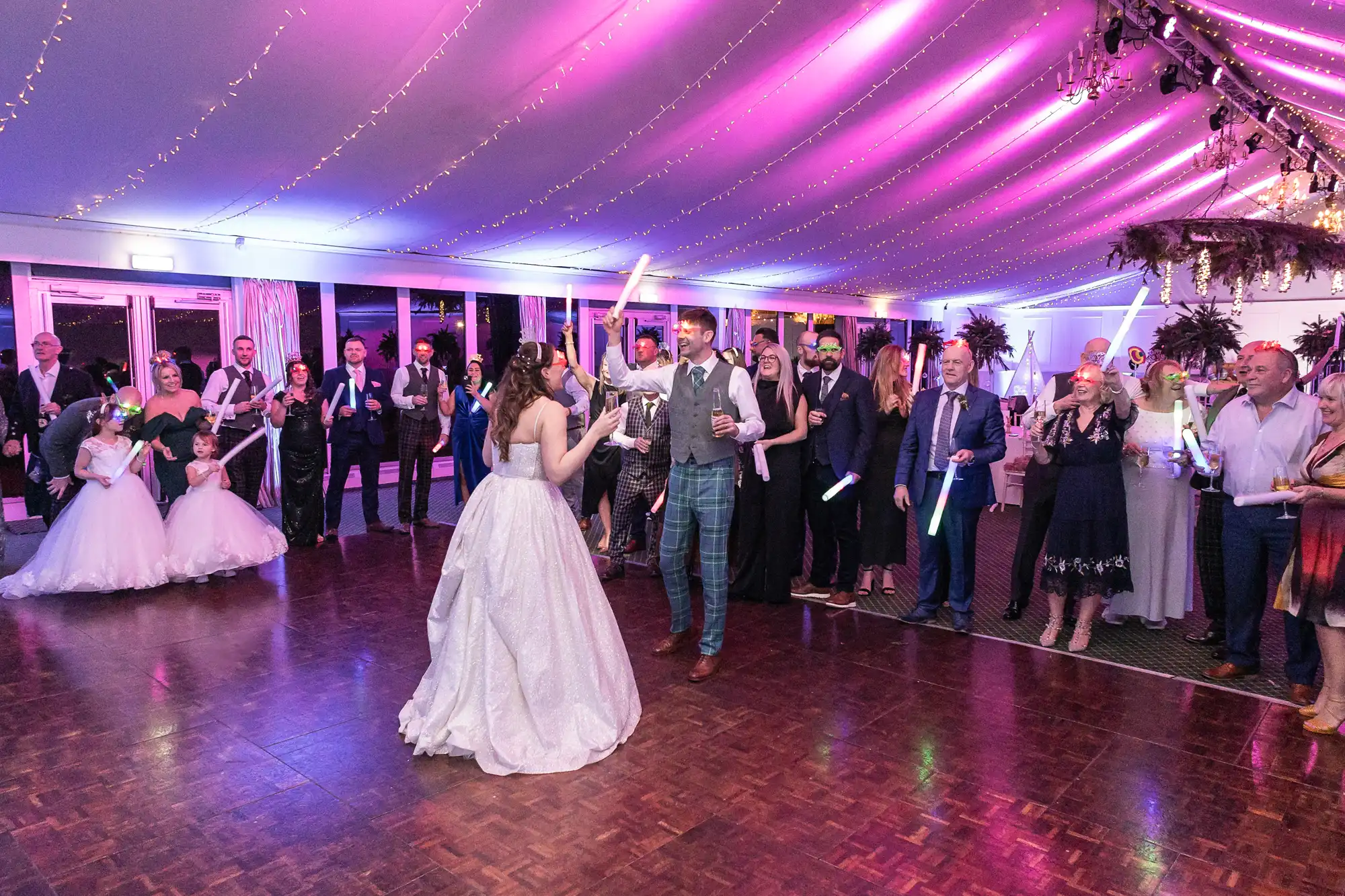 A bride and groom dance under purple lighting in a tent, surrounded by guests holding glow sticks.