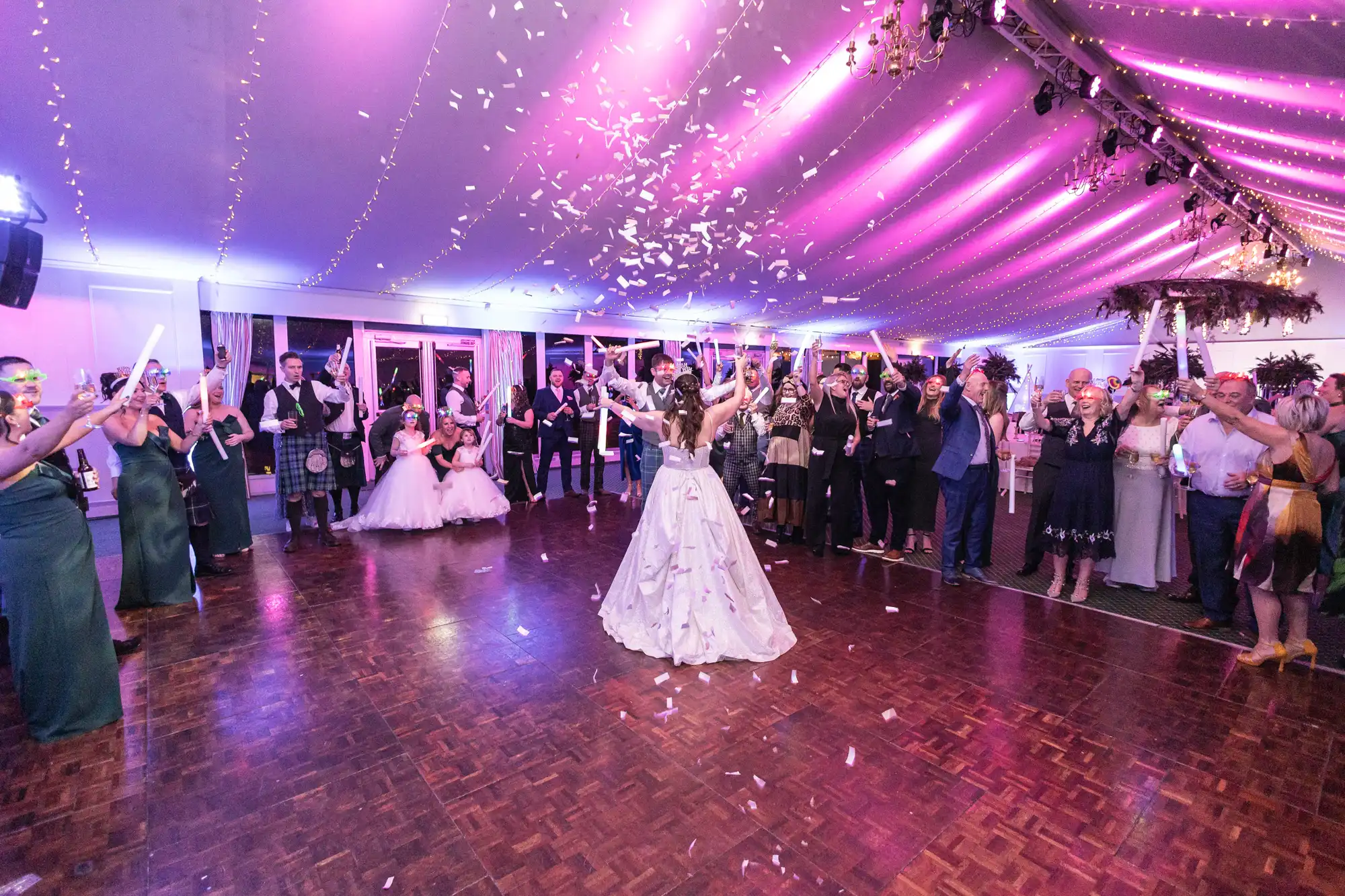 A bride stands in the center of a dance floor with guests surrounding her in celebration. Confetti fills the air under pink ceiling lights in a decorated reception venue.