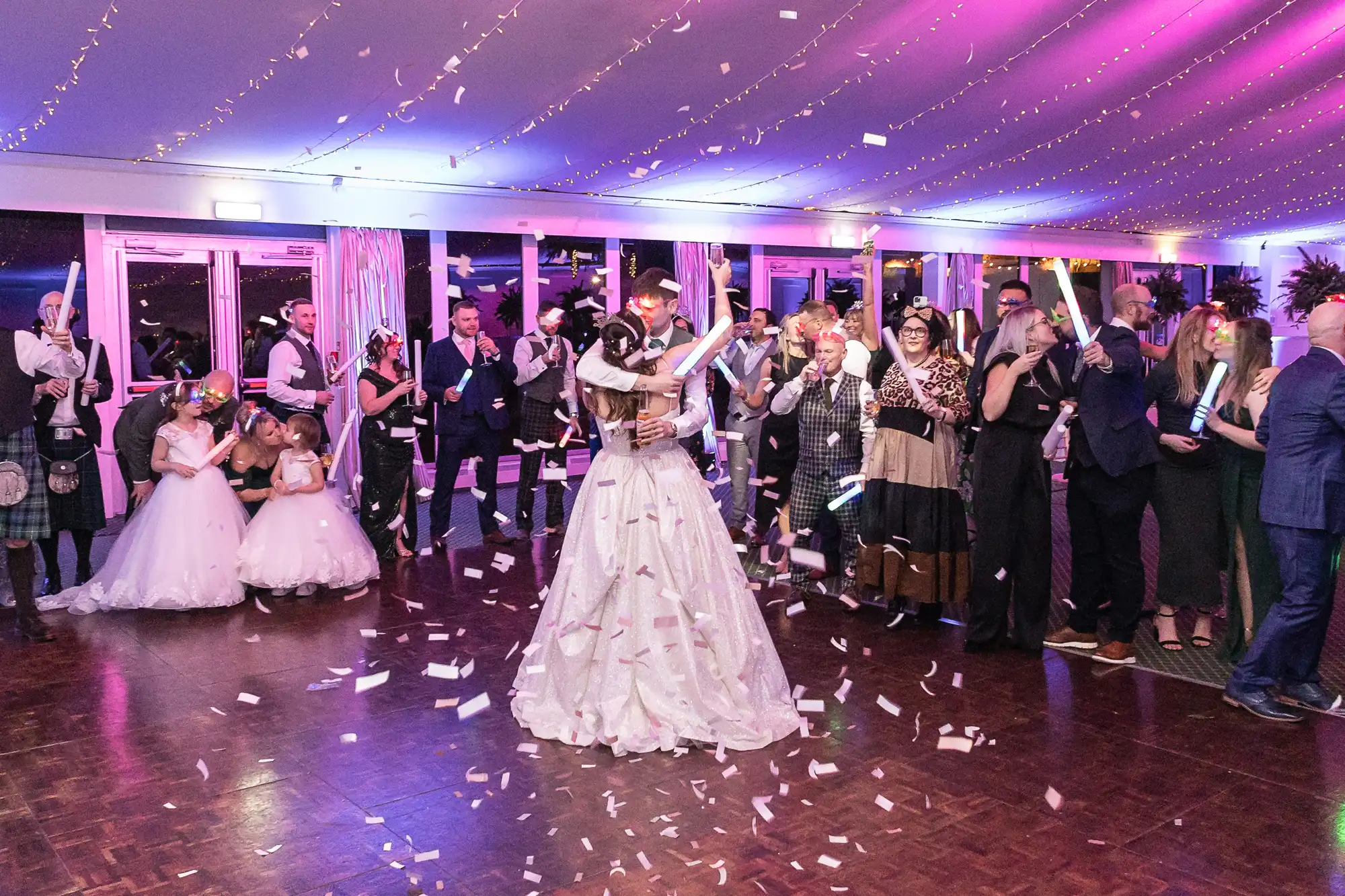 A couple dances in the center of a confetti-filled dance floor, surrounded by guests dressed in formal attire under purple and blue lighting.