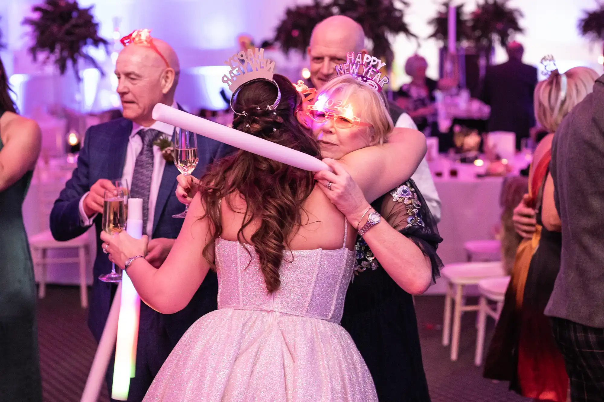 People celebrating New Year's Eve at a party, wearing festive hats and glasses. A woman in a white dress is hugging another woman while holding a champagne flute.