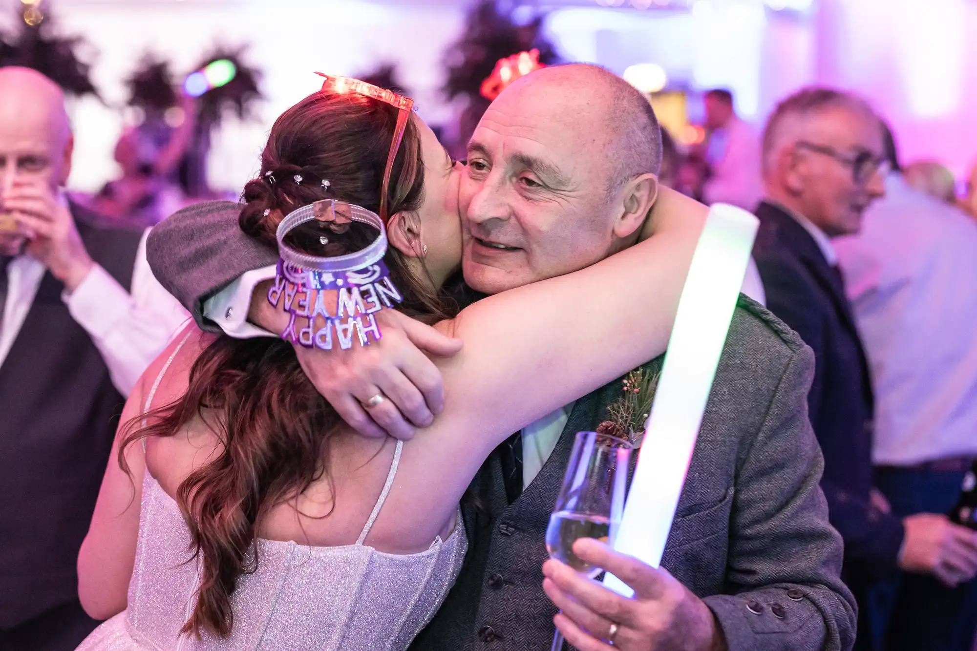 A woman and an older man hug at a celebratory event, both holding glowing light sticks. The woman wears a "Happy New Year" headband.