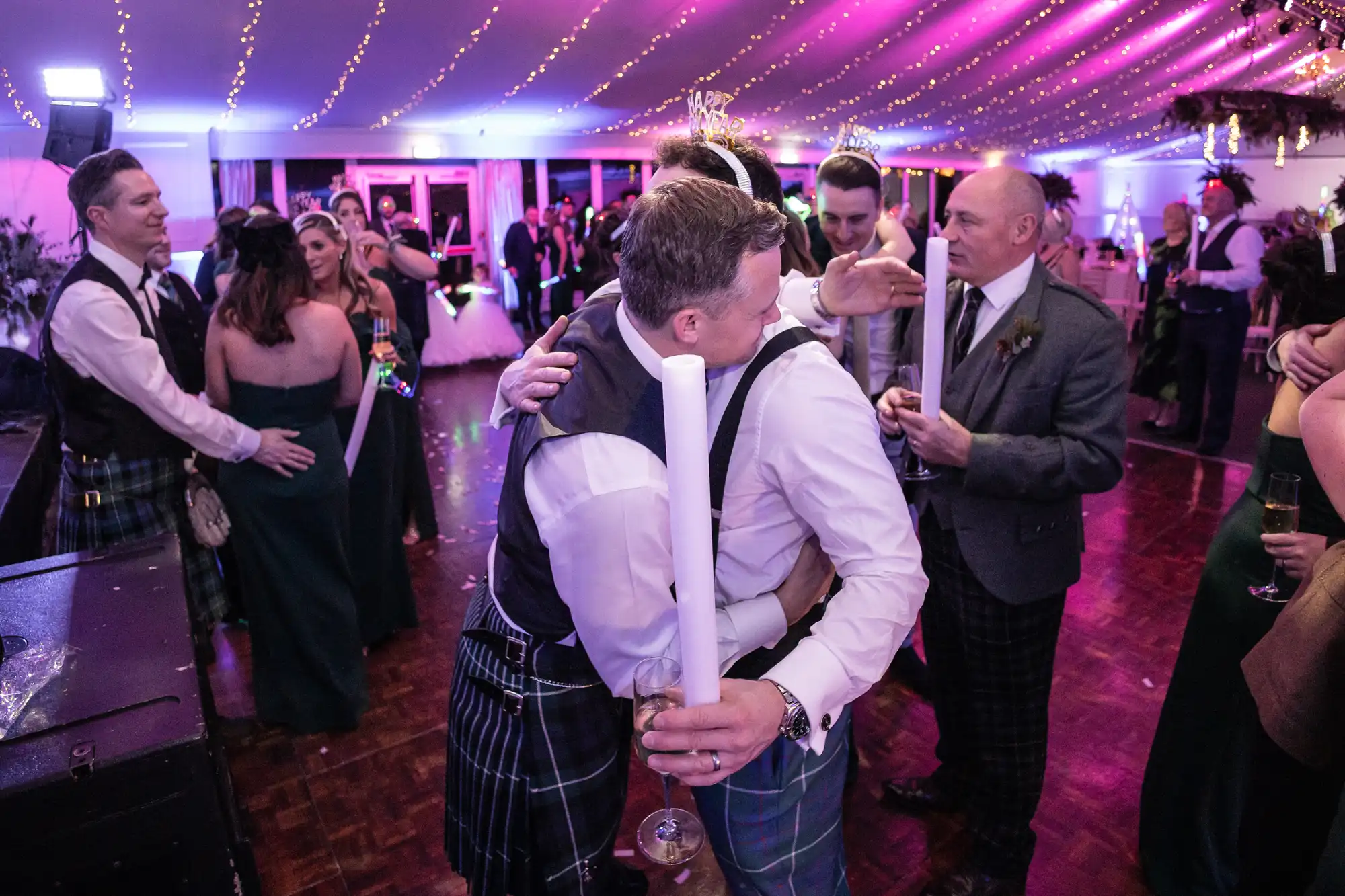 A group of people at a festive event, some wearing kilts, are gathered closely together under purple string lights. Two men are hugging in the center while others are engaged in conversation.